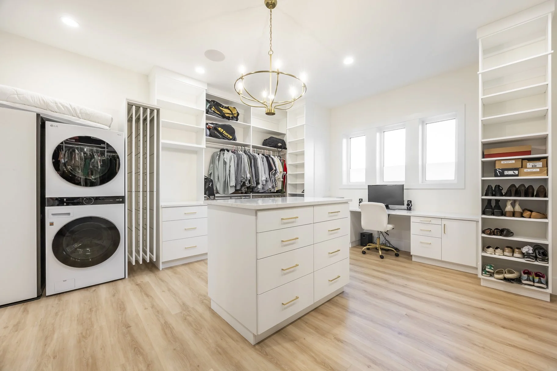 Bright laundry room with white walls and cabinets, wooden floor, washer and dryer stacked on the left, clothing and shoes stored on shelves, a small desk with a computer near windows, and a gold chandelier hanging from the ceiling.