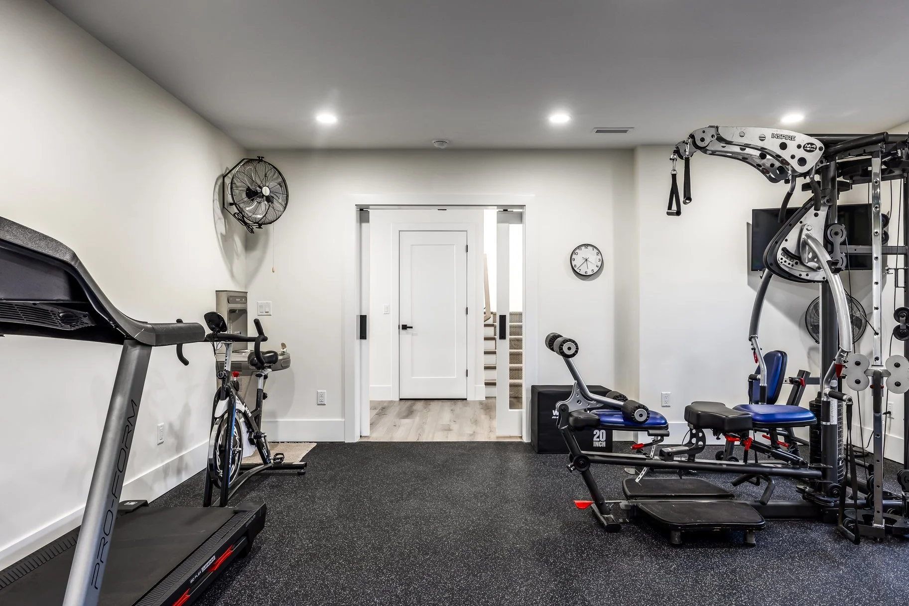 empty home gym with treadmill, exercise bike, weight machine, and equipment, white walls, ceiling fans, clock, and stairs in background