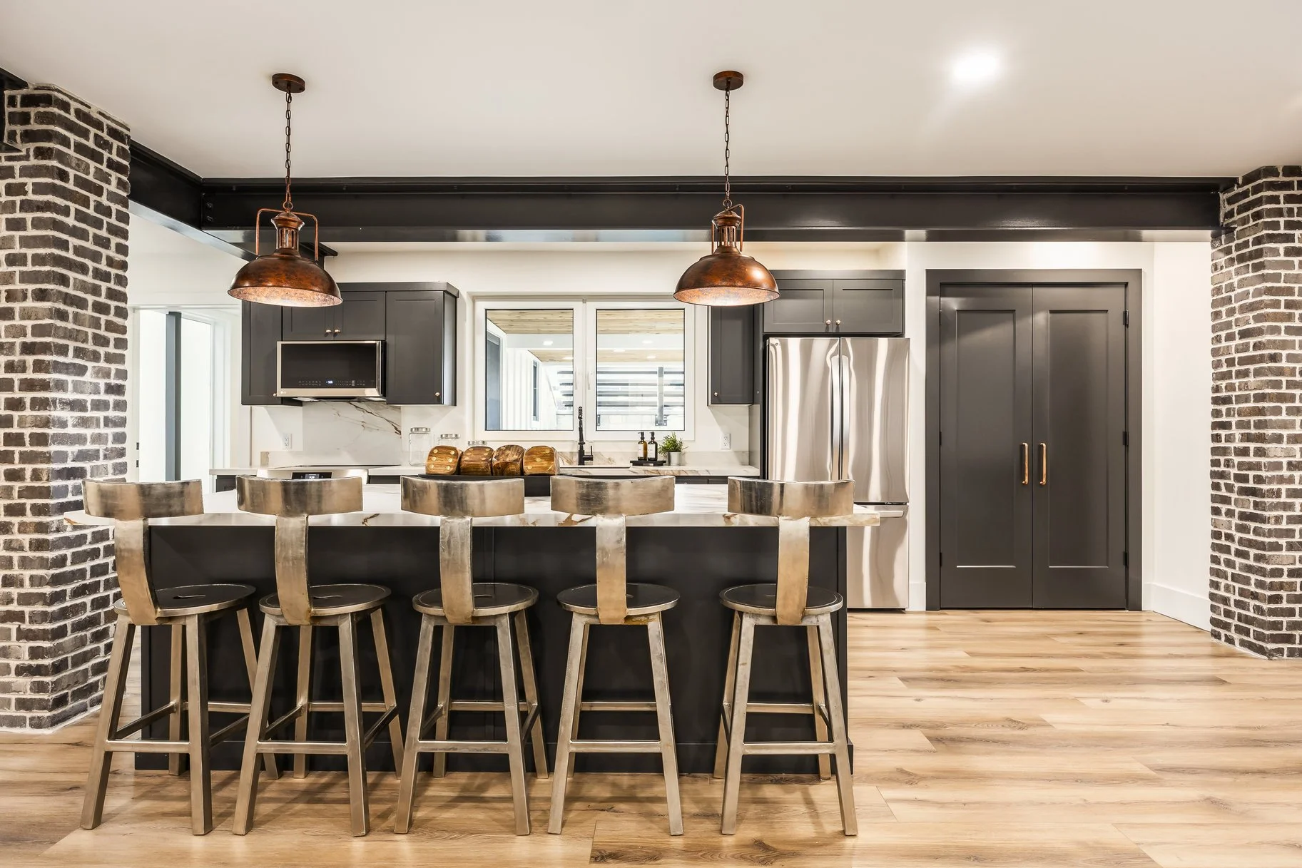 Modern kitchen with black cabinets, stainless steel appliances, brick columns, wooden barstools, and pendant lighting.