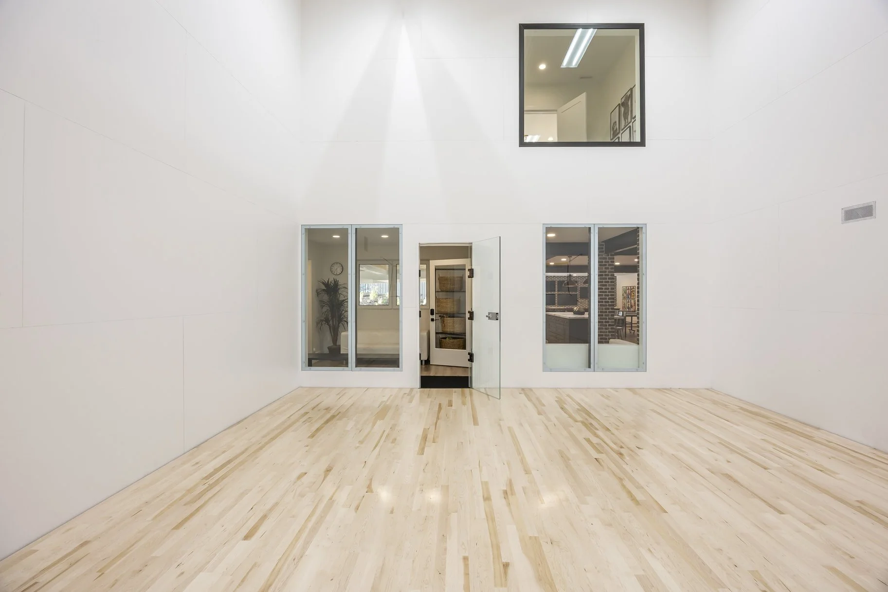 Empty room with light wood flooring, white walls, and a large black-framed window. There are glass doors and windows leading to another room with a plant, clock, and visible kitchen area. A smaller window is present near the ceiling.