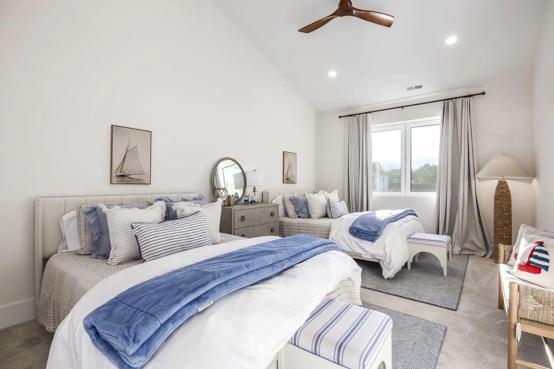A bright bedroom with two beds, each with white and blue striped bedding, pillows, and blankets. There is a window with beige curtains, a bedside table with a lamp, a dresser with a round mirror, a ceiling fan, and a cozy seating area with a wicker c