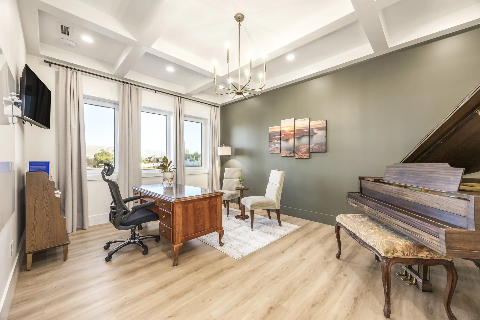 Bright, spacious home office with light wooden flooring, a wooden desk with a black office chair, two white armchairs, a piano, a wall art piece, and windows with beige curtains.