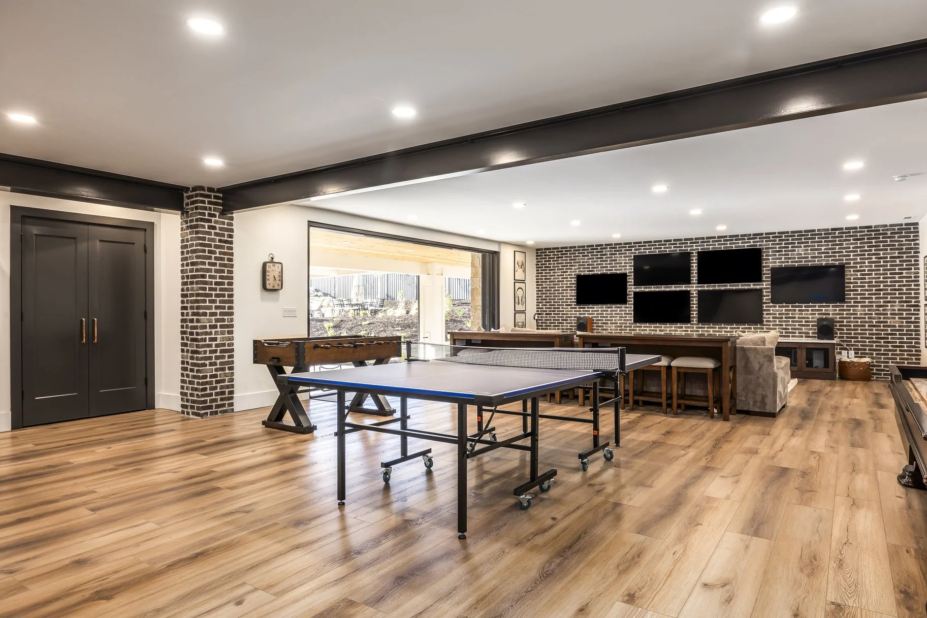 A spacious entertainment room with a ping pong table, a foosball table, and a bar area with multiple TVs on a brick wall.