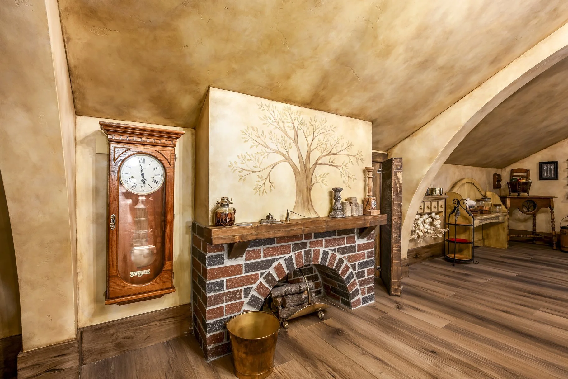 A cozy room with a brick fireplace and a wooden mantel, decorated with small jars and candlesticks. Above the fireplace is a wall painting of a large tree, and next to it is a vintage wooden grandfather clock. The room features wooden floors, an arch