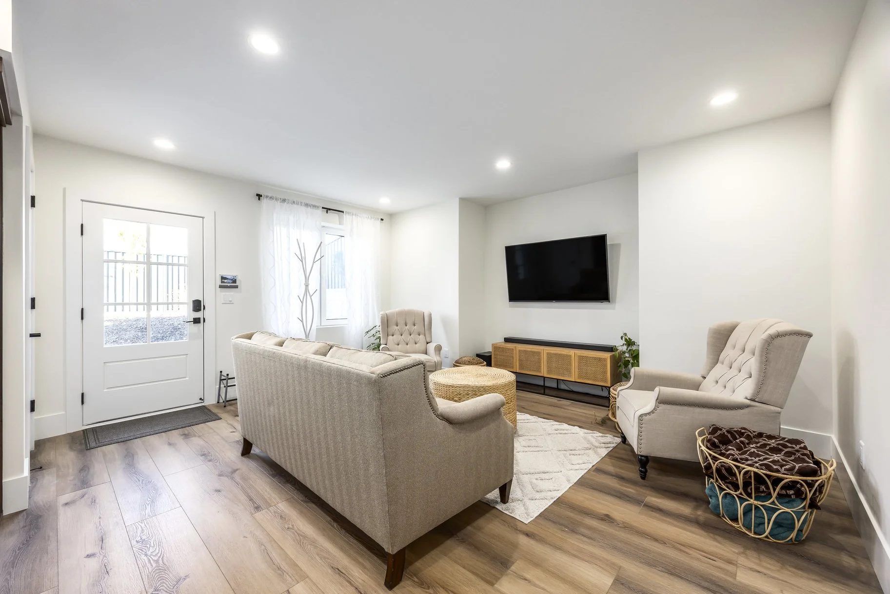 Living room with beige sofa, armchairs, a wall-mounted TV, a wooden TV stand, a rug, wicker basket, and large windows with white curtains.