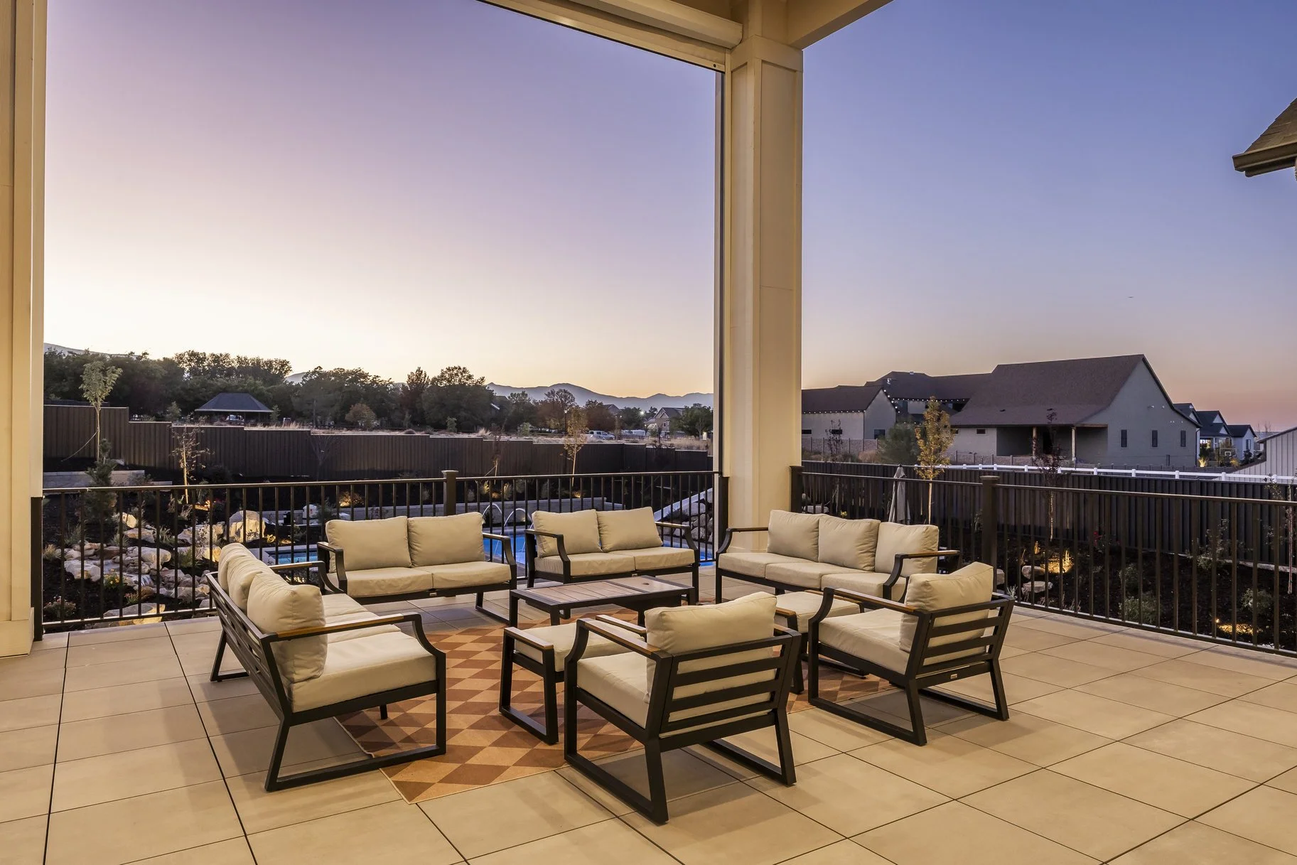 An outdoor patio with six cushioned chairs arranged around a small table on a patchwork rug, overlooking a neighborhood at sunset with houses, trees, and mountains in the background.