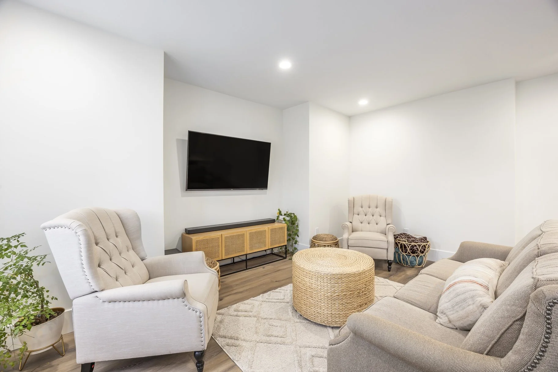 Contemporary living room with beige upholstered armchairs, a beige sofa, a flat-screen TV on the wall, a wooden media console, a woven round ottoman, a patterned rug, and houseplants.