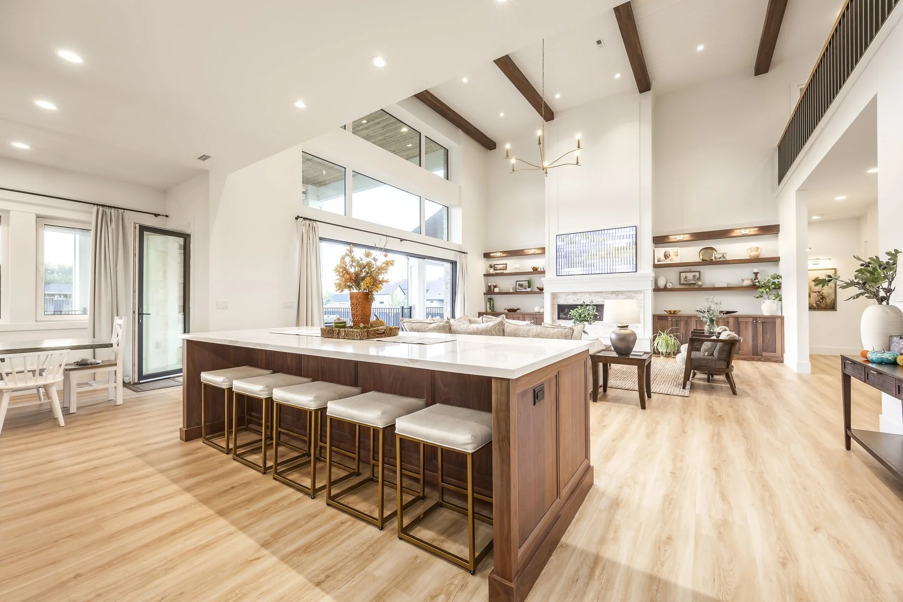 Open-concept living room and kitchen with high ceilings, wooden beams, a large island with four barstools, and a seating area with a fireplace and fireplace mantel. Light-colored hardwood flooring and decorative shelves with plants and ornaments.