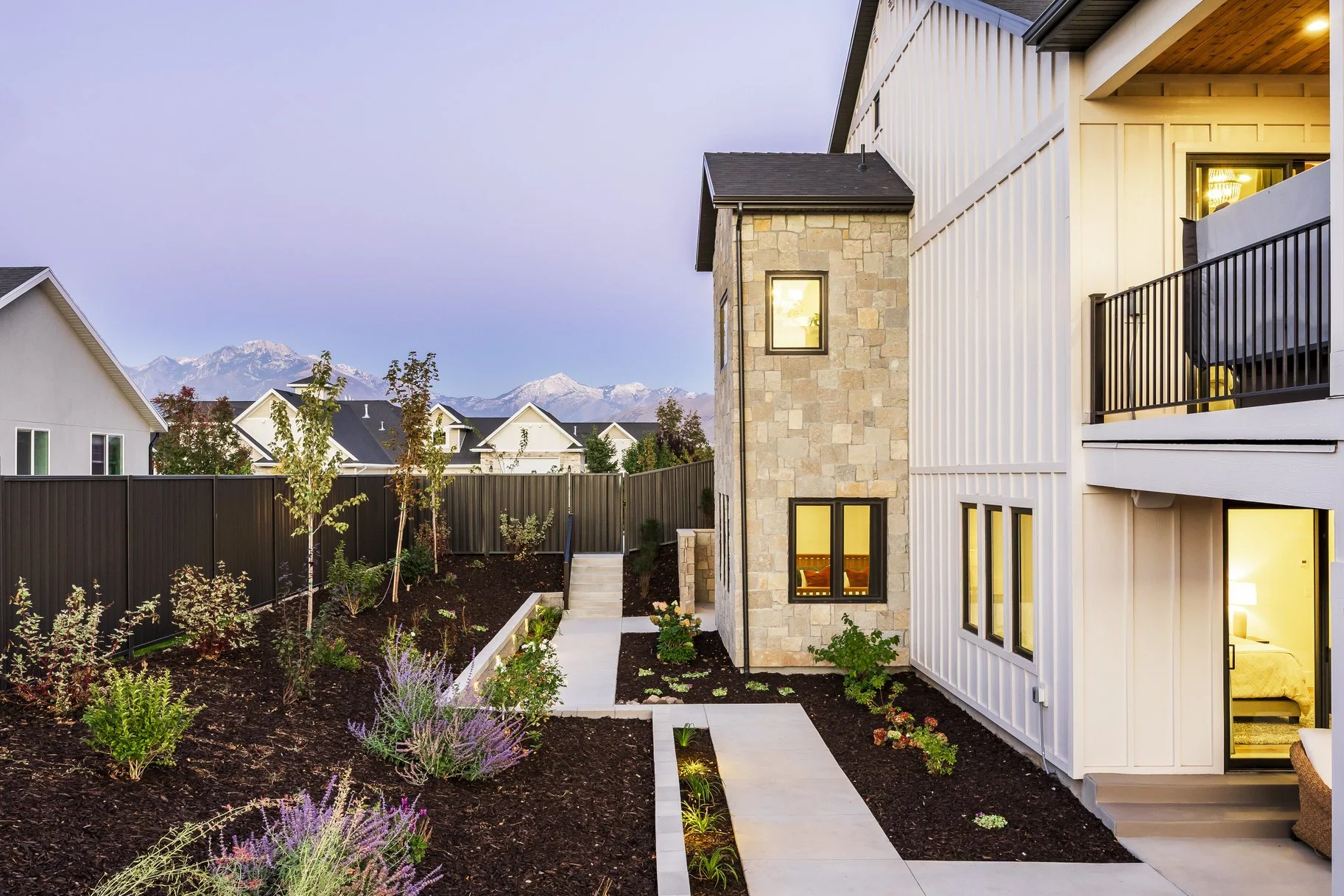View of a modern residential backyard with a paved walkway, landscaped garden, and a multi-story house with a stone wall, white siding, and large windows, with mountains in the background.