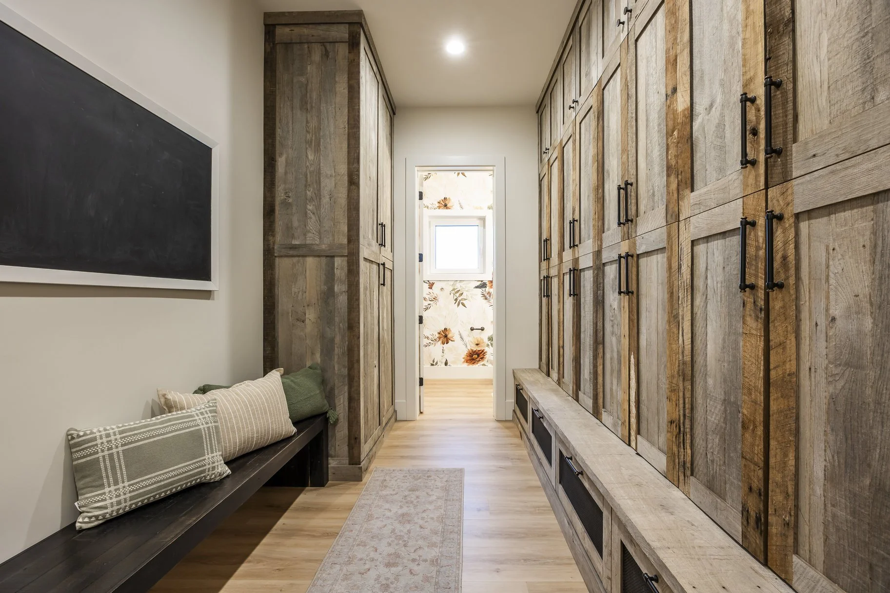 A hallway with wooden storage cabinets on the right, a bench with pillows on the left, a blackboard on the wall, hardwood floors, a small rug, and a doorway at the end with floral wallpaper and a window.