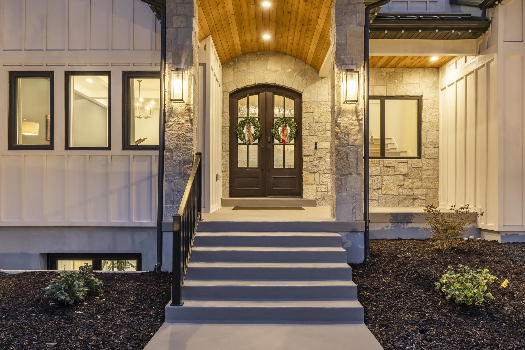 Front entrance of a house with double wooden doors decorated with wreaths, stone and white siding exterior, steps leading up to the door, and warm lighting.