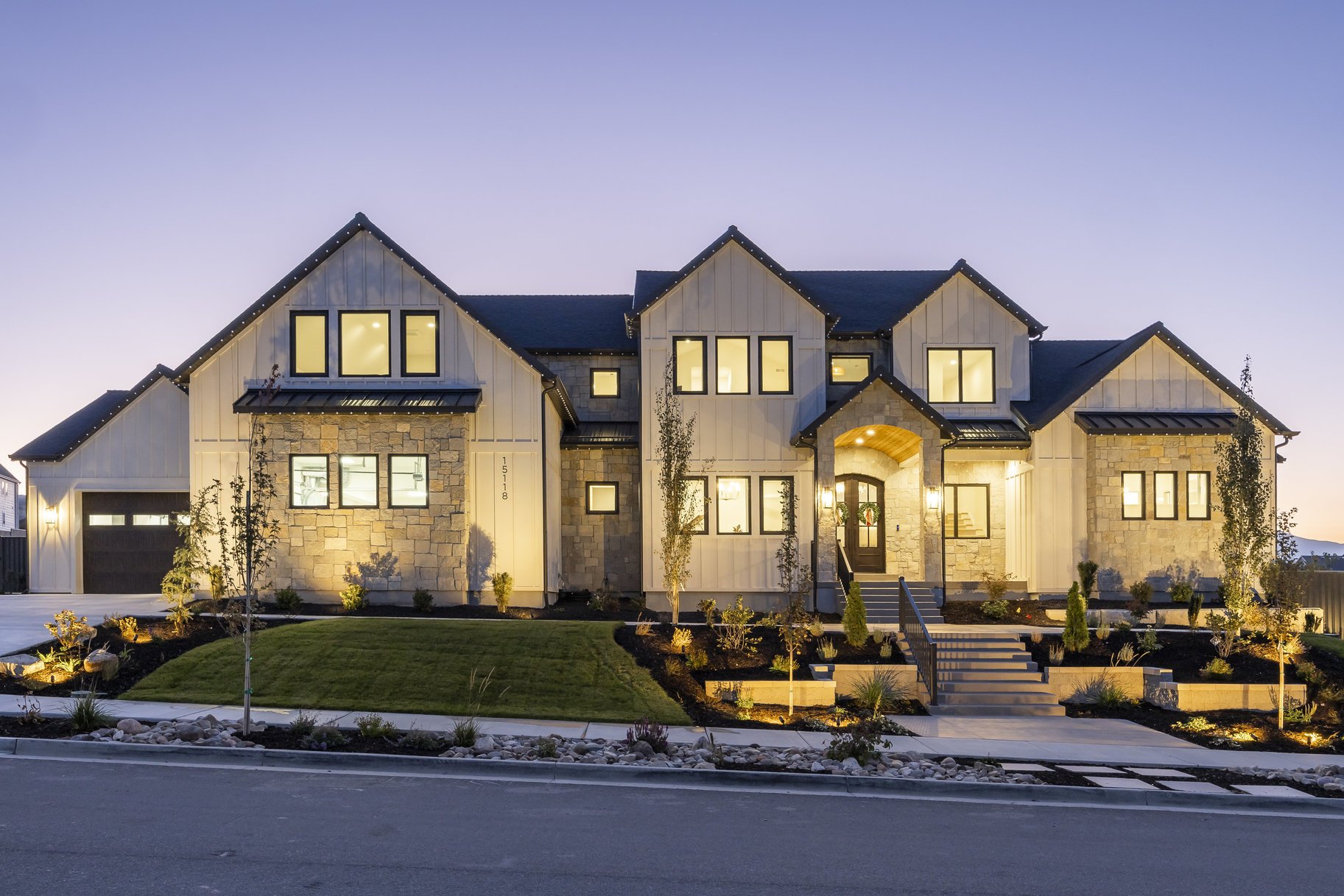 Modern two-story house with stone and light-colored siding, illuminated at dusk, featuring a well-manicured lawn, young trees, and landscaped front yard.