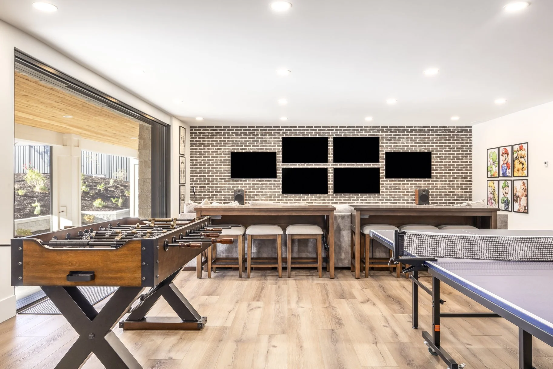 A game room featuring a foosball table, ping pong table, and seating area with five black TVs mounted on a brick wall.
