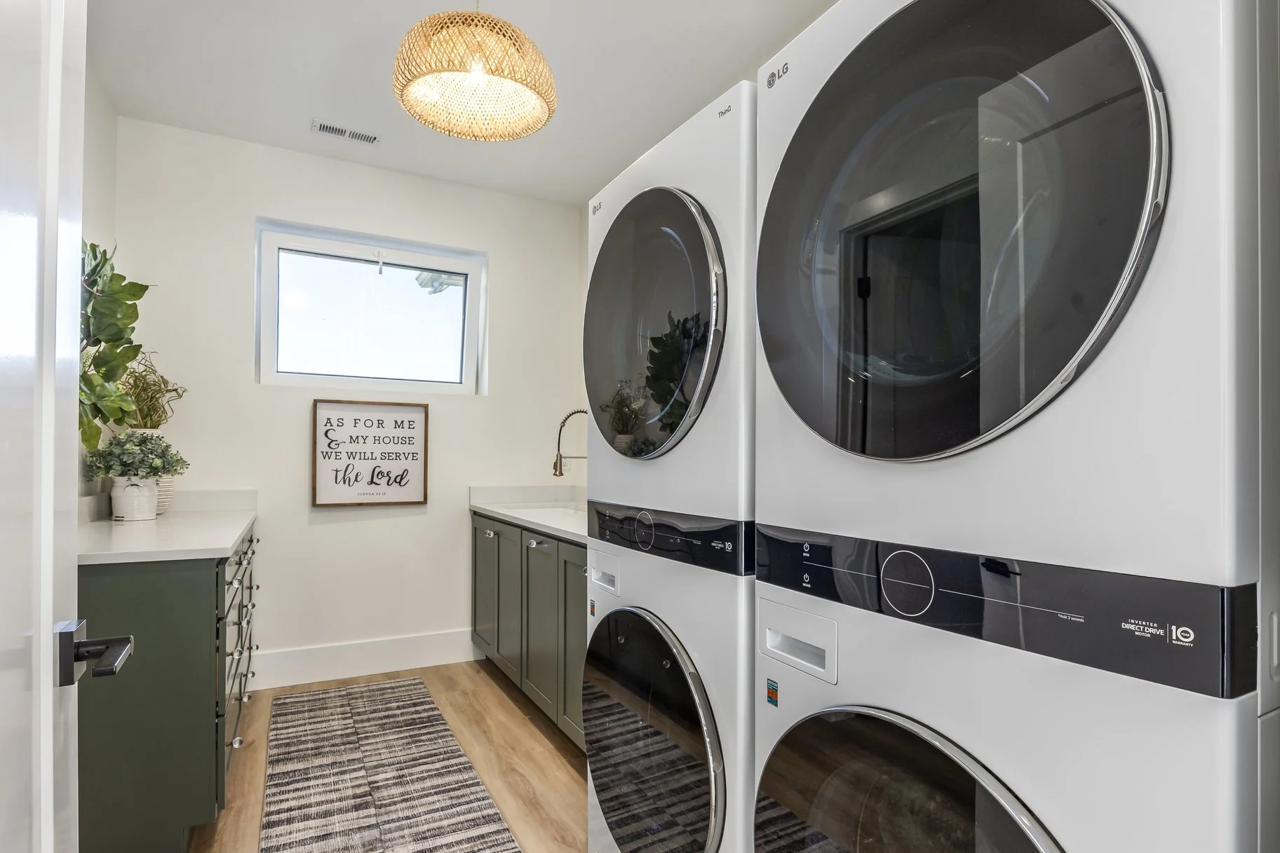 A laundry room with a stacked washer and dryer next to a sink, cabinets, a window, a framed sign, plants, a hanging light fixture, and a rug.