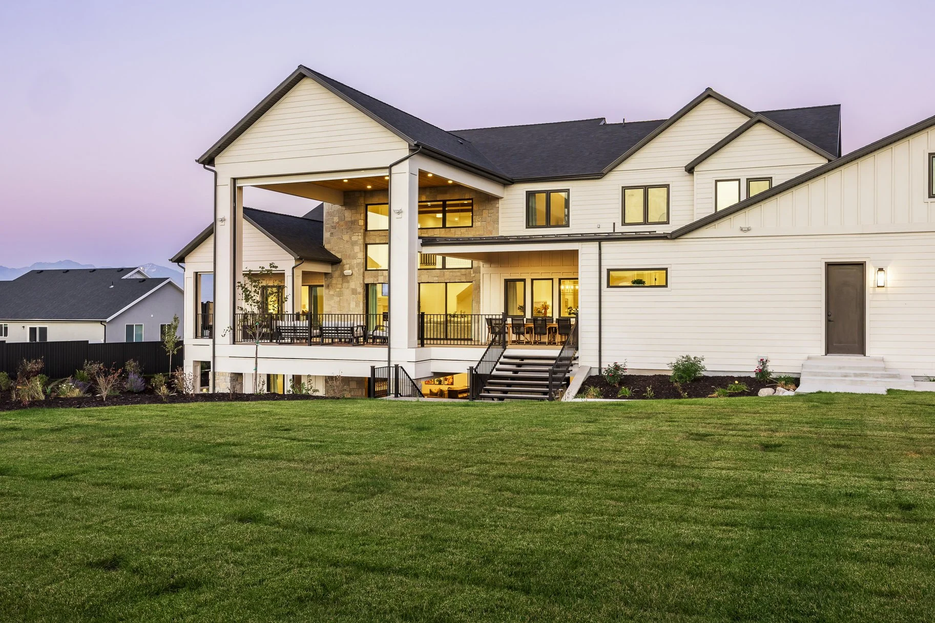 Modern two-story house with large balcony and outdoor seating, white siding, black roof, and landscaped lawn