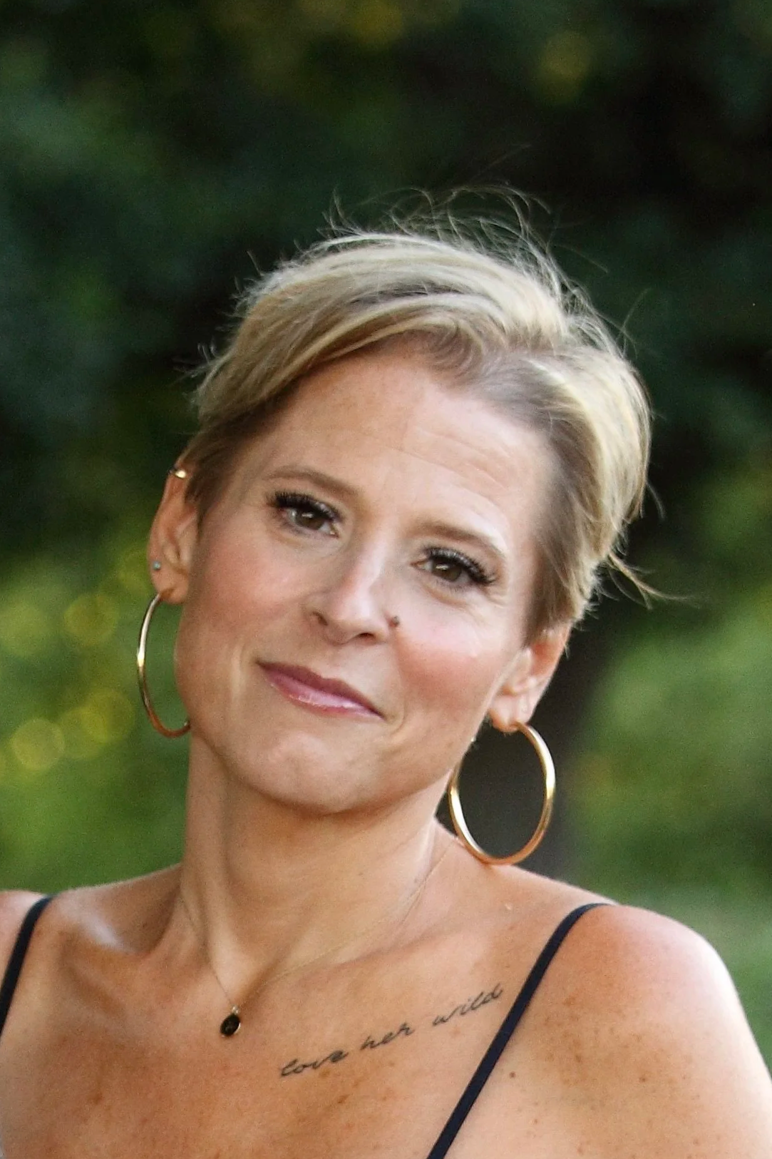 A woman with short blonde hair posing outdoors, wearing large hoop earrings and a black tank top, with a tattoo that reads 'love her wild' on her chest, and a small necklace.