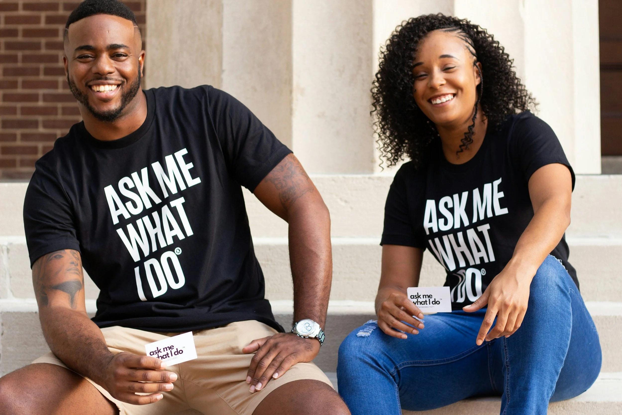 A smiling man and woman sitting on steps outdoors, holding small cards that say "ask me what I do". Both are wearing black T-shirts with the same phrase printed on them.