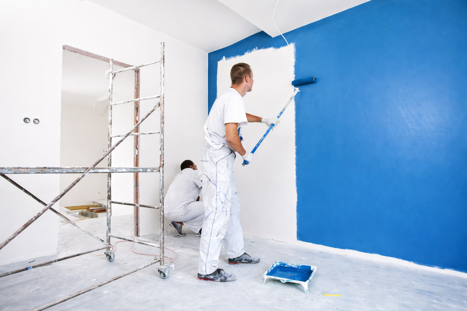 Two painters painting a blue wall in a room, with one standing and using a roller, and the other crouched on the floor nearby.