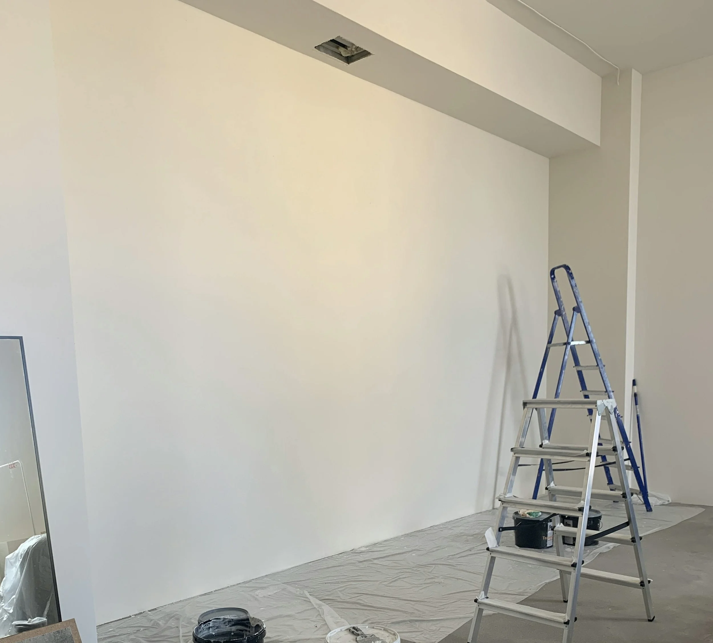 Empty room with painting supplies, including a ladder, paint buckets, and plastic covering on the floor, indicating ongoing interior painting or renovation.