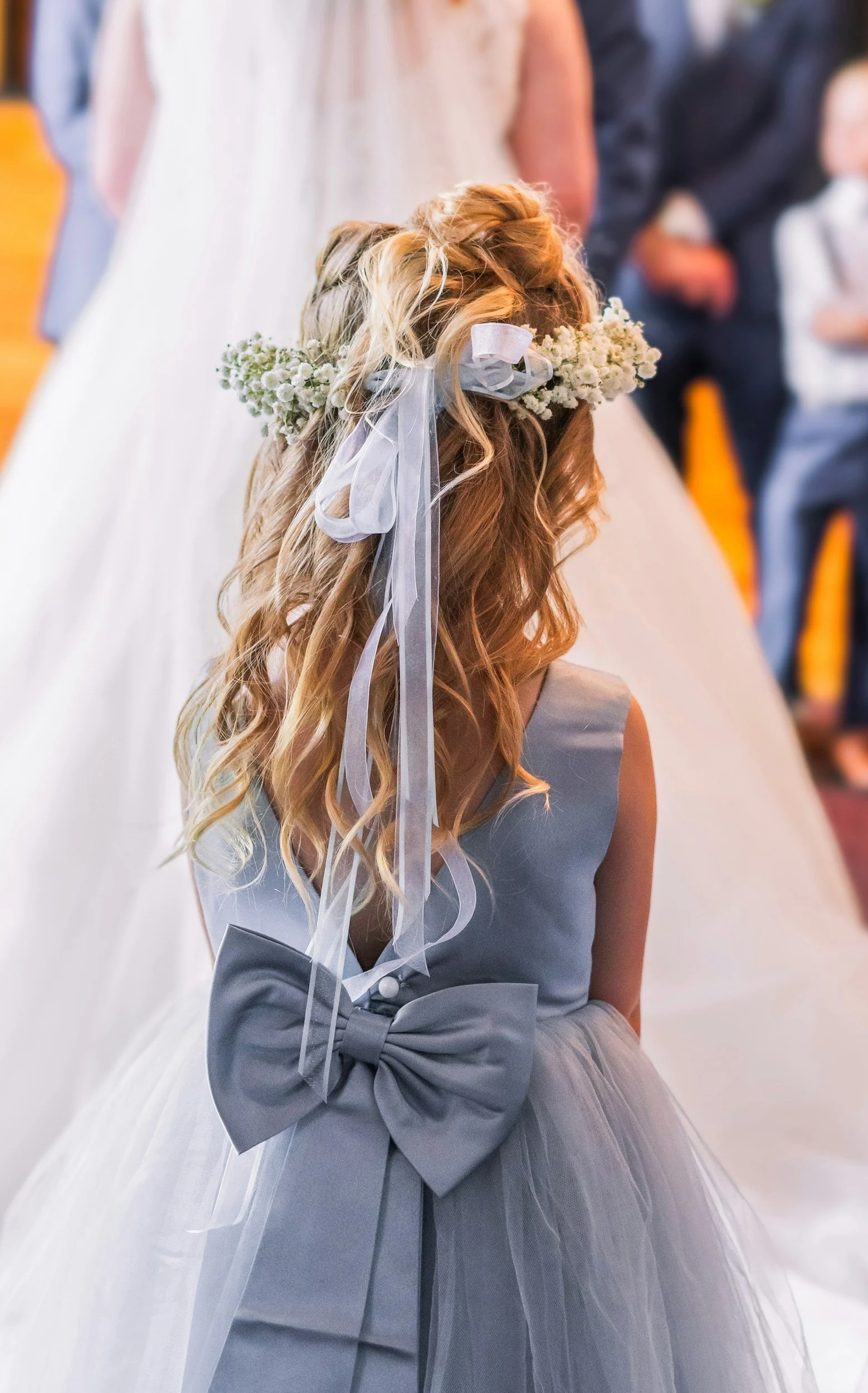 A flower girl standing in a wedding. The back of her head is shown, along with her hair piece and details of her dress.