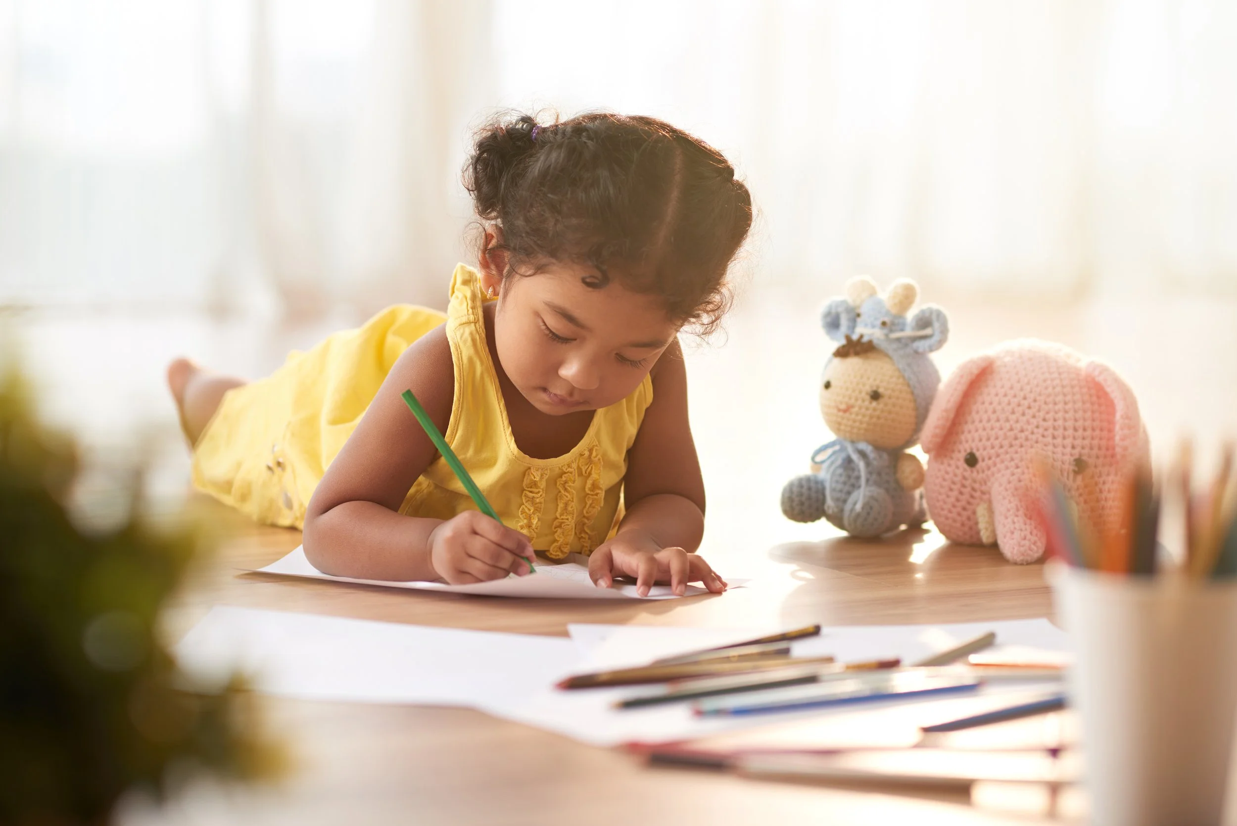 Young girl in a yellow shirt laying on the floor coloring with colored pencils