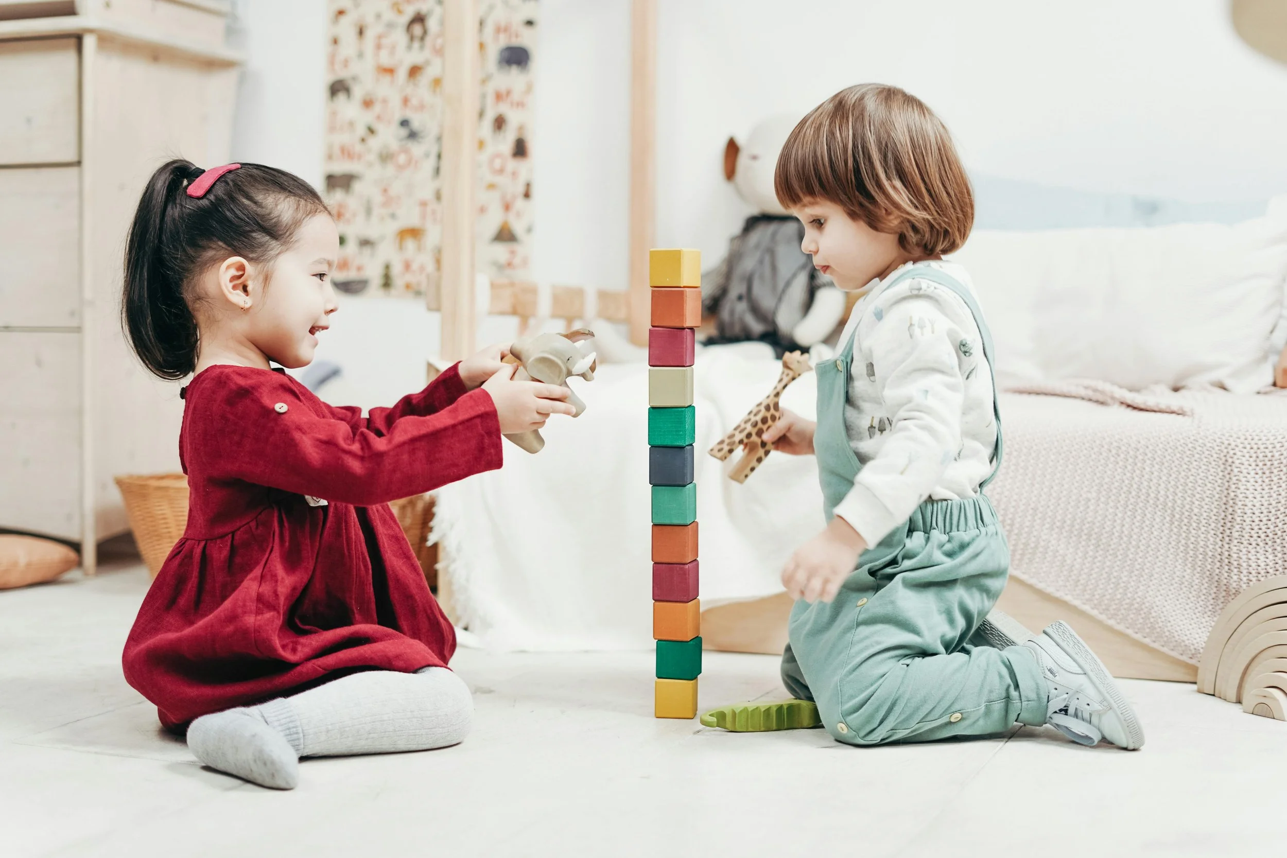 A girl in a red dress and a boy in blue overalls playing with toys. She is holding an elephant and he is holding a giraffe. Between them is a tower of colorful wooden blocks.