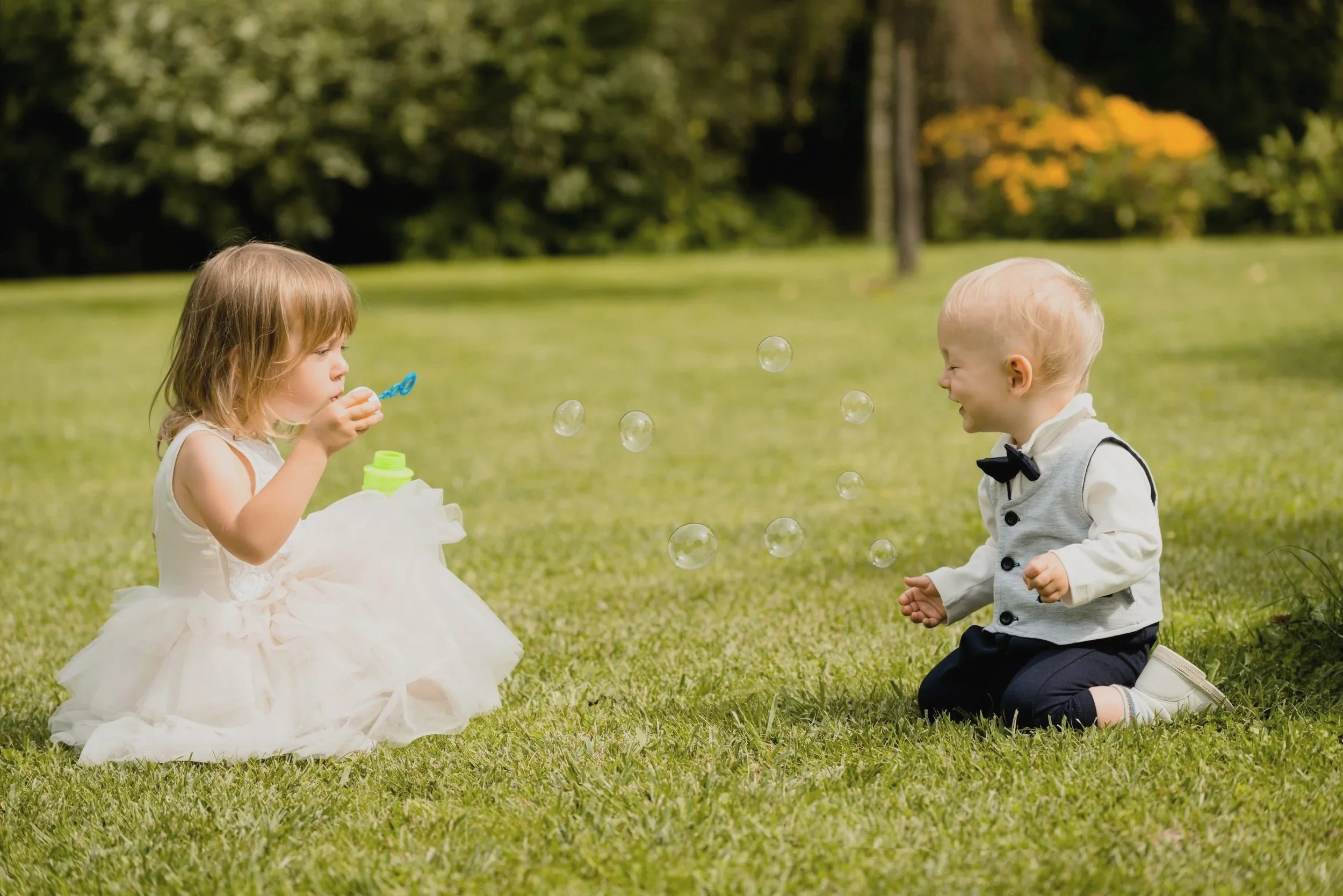 Flower girl and another child playing with bubbles while sitting in the grass at a wedding