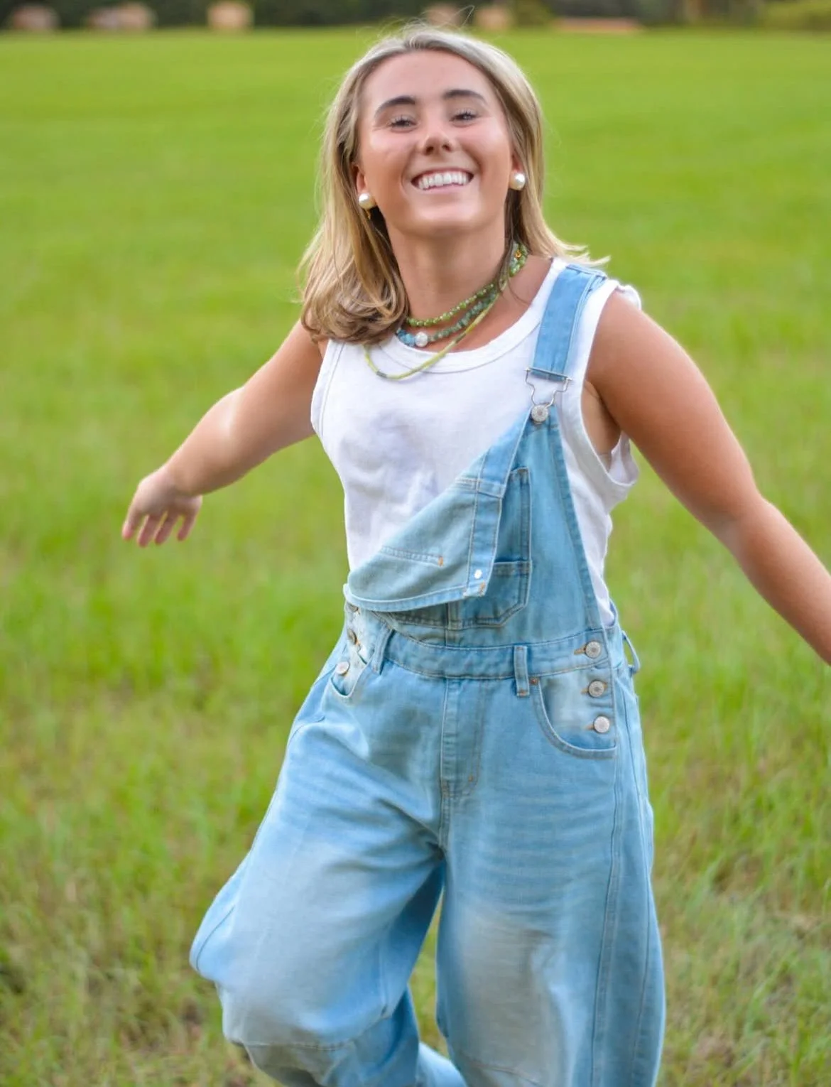 A woman in denim overalls and a white tank top standing in a grassy field with arms outstretched, smiling with her hair flowing, during daytime.