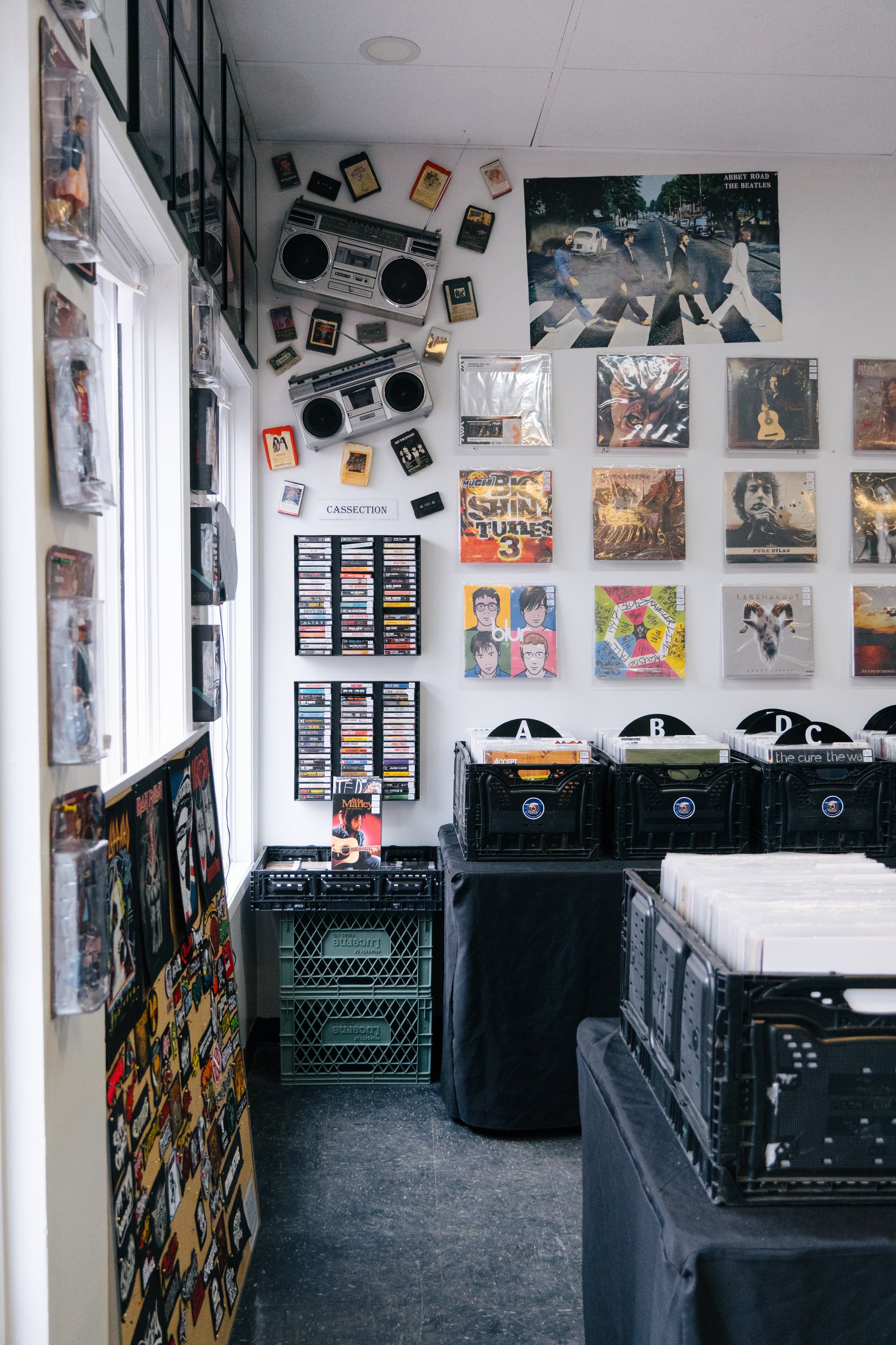A room with vinyl records, cassette tapes, and music-related posters on the walls, including a large poster of The Beatles crossing Abbey Road. There are black crates filled with records on tables covered with black cloths.