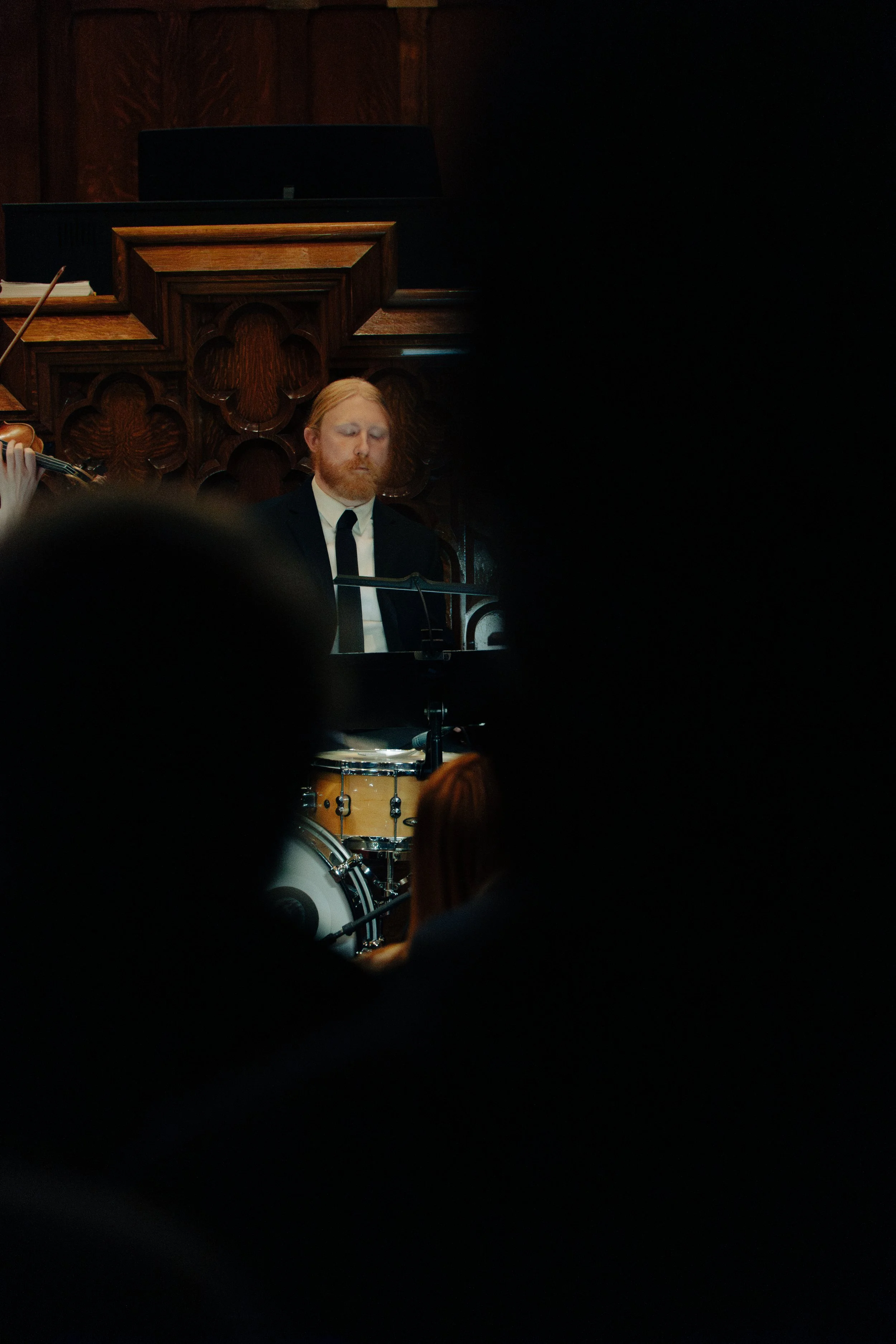 A man with red hair and a beard playing the piano during a musical performance, with a drum in the foreground and a wooden wall in the background.