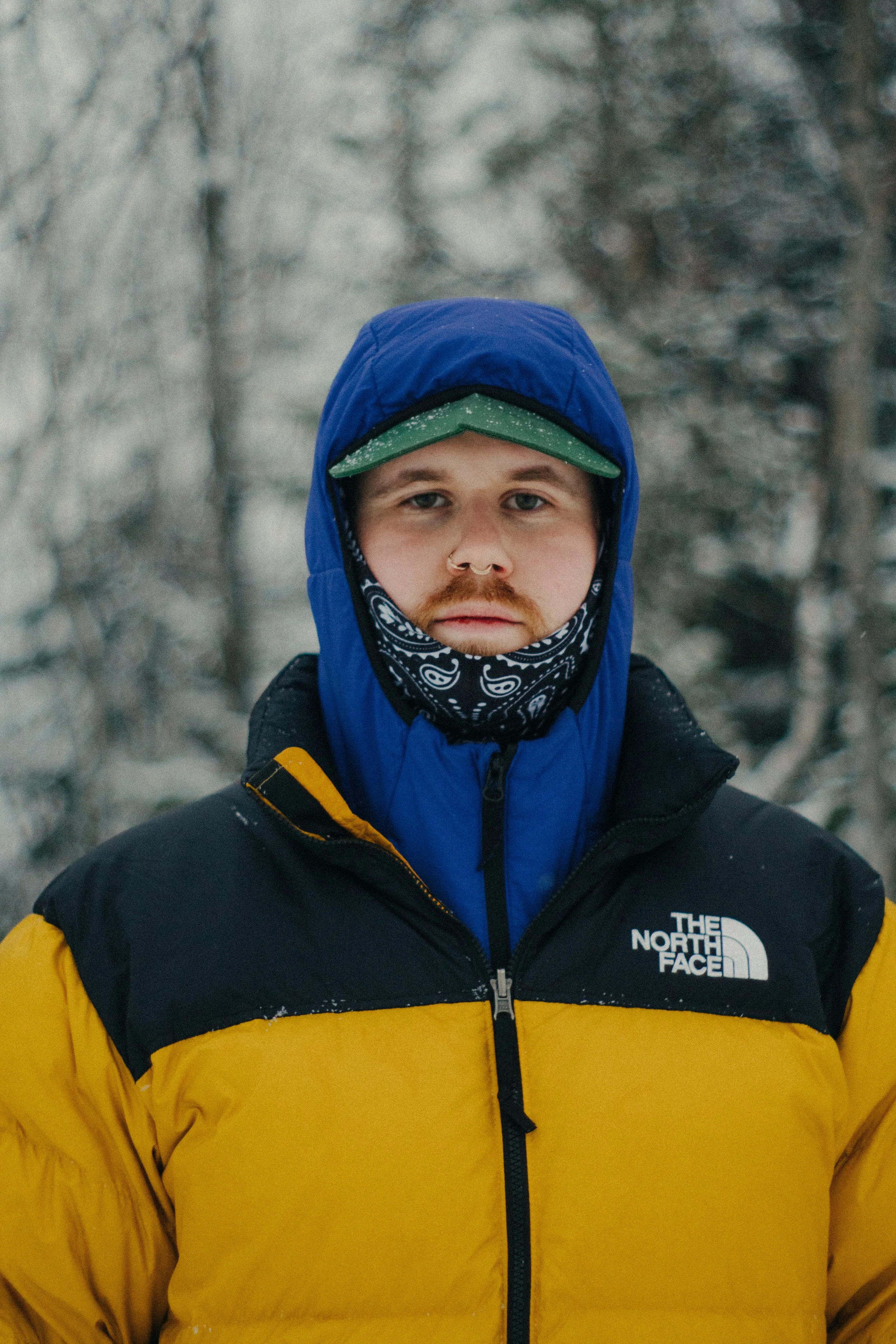 A man in outdoor winter clothing, including a blue hoodie over a green cap, a bandana mask, and a yellow and black jacket with 'The North Face' logo, standing outdoors in a snowy, wooded area.