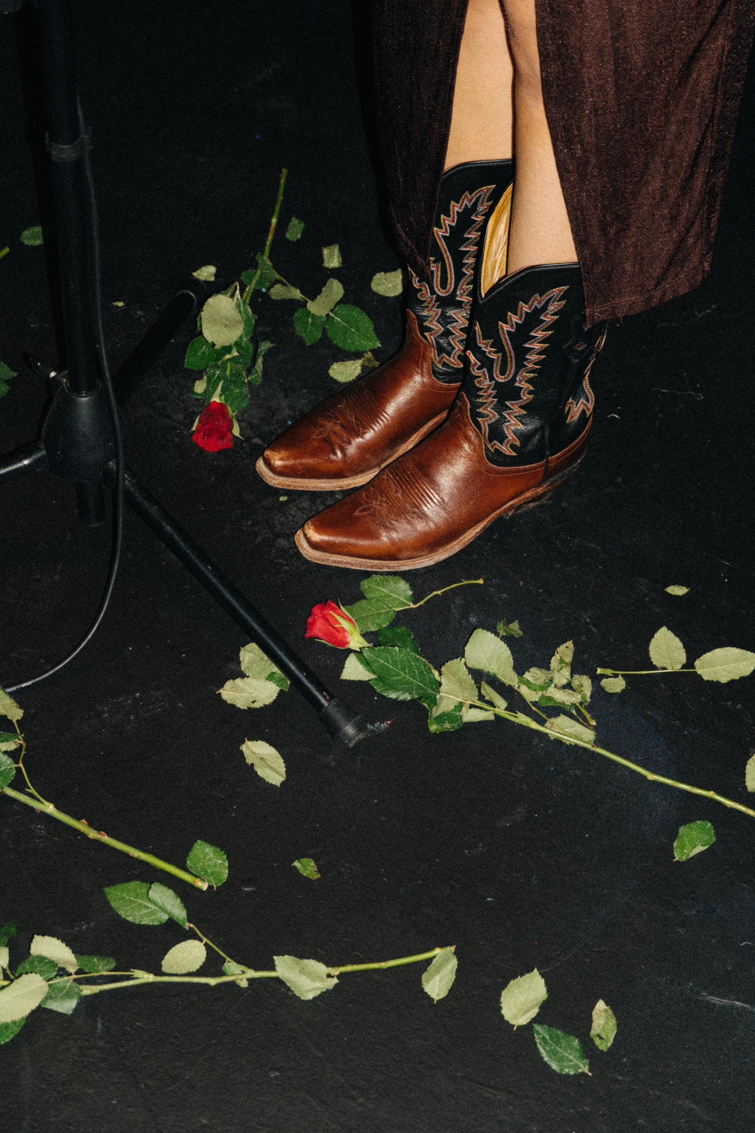 A person's lower legs and feet wearing cowboy boots with a black and colorful embroidered design, dark pants, and a long brown coat, standing on a black floor surrounded by scattered green and yellow leaves and a red rose.