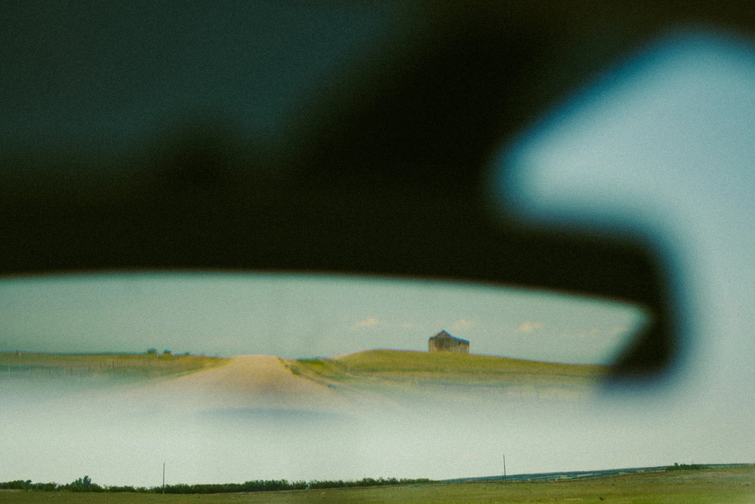 A rural landscape with a single house in the distance under a blue sky, viewed through a car window with a blurred border in the foreground.