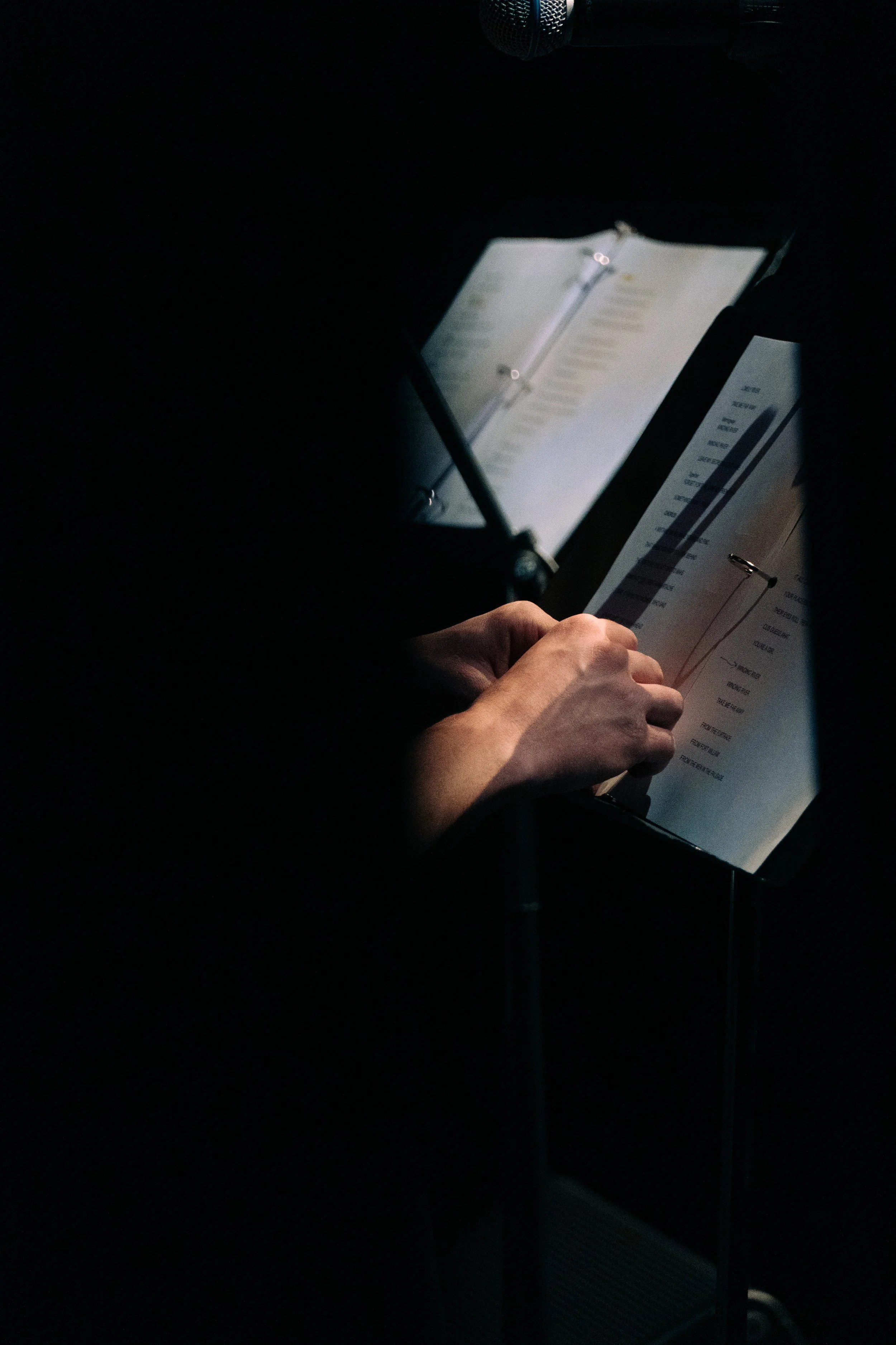 A person's hand is turning a page of a music sheet on a stand, with a microphone above in a dark setting.