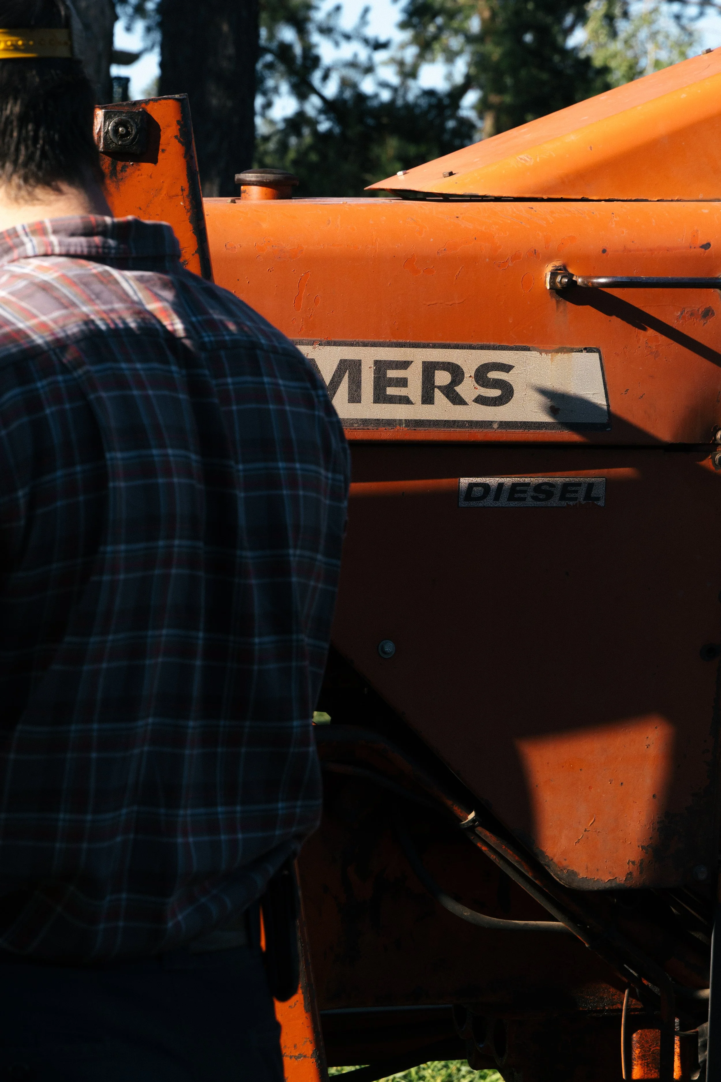 Close-up of a person in a plaid shirt near an orange industrial machine, with visible signs of wear and a shadow cast on the machine.