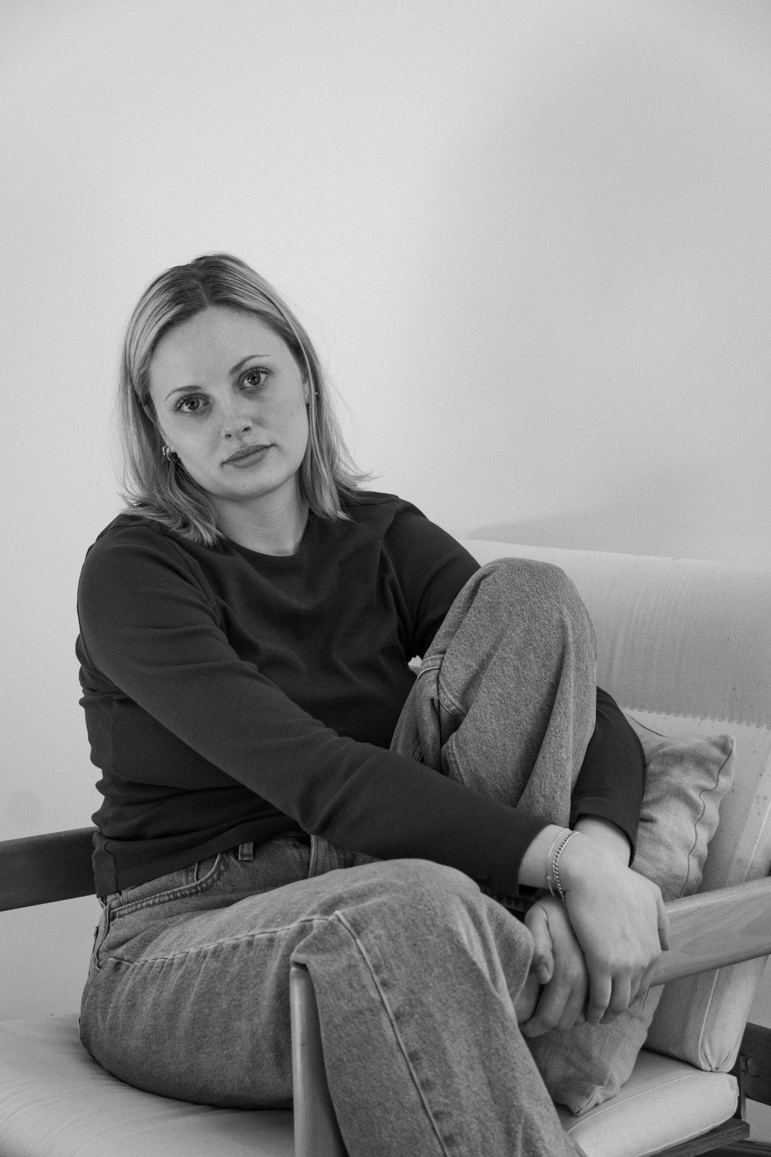 Black and white photo of a woman with shoulder-length hair, sitting on a bed with a plain wall in the background. She is wearing a long-sleeve shirt and jeans, with a serious expression on her face, holding her sleeve with her hands.