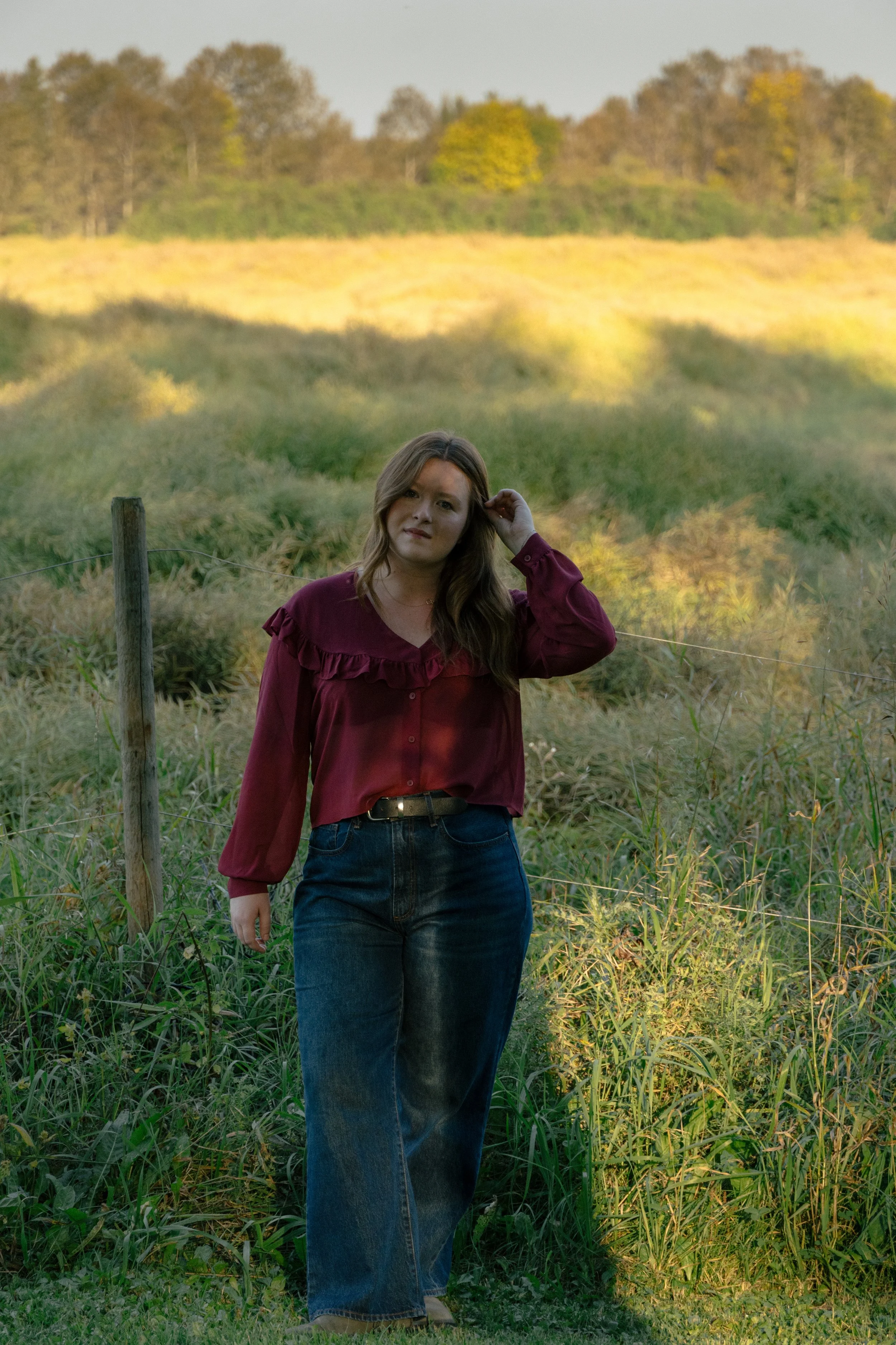 A woman with brown hair wearing a burgundy blouse and blue jeans standing in a grassy field with a fence and a backdrop of trees and rolling hills.