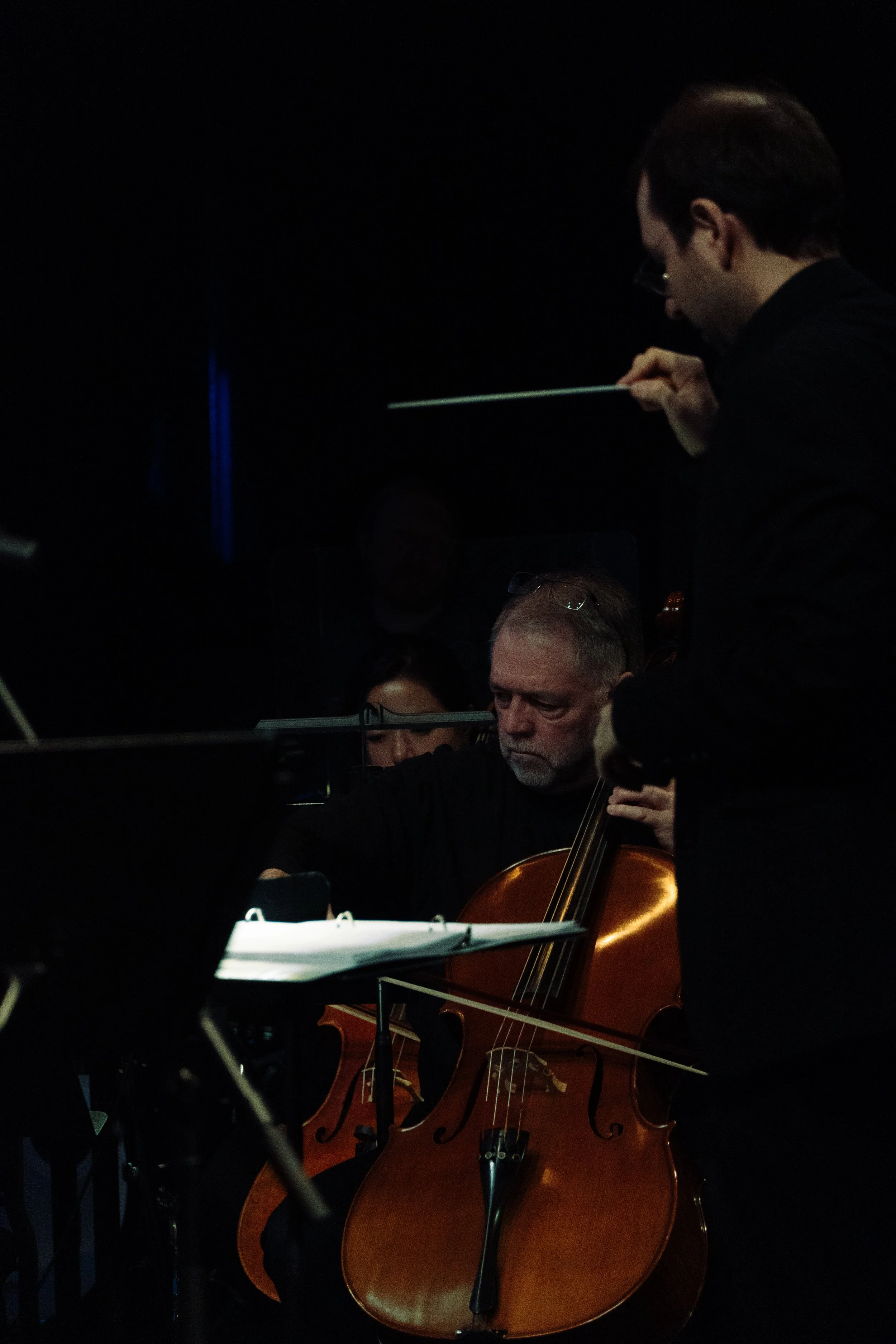 Musicians performing in a dark concert hall: a man with glasses playing the cello, and a conductor with short brown hair, glasses, and a black suit leading the performance.