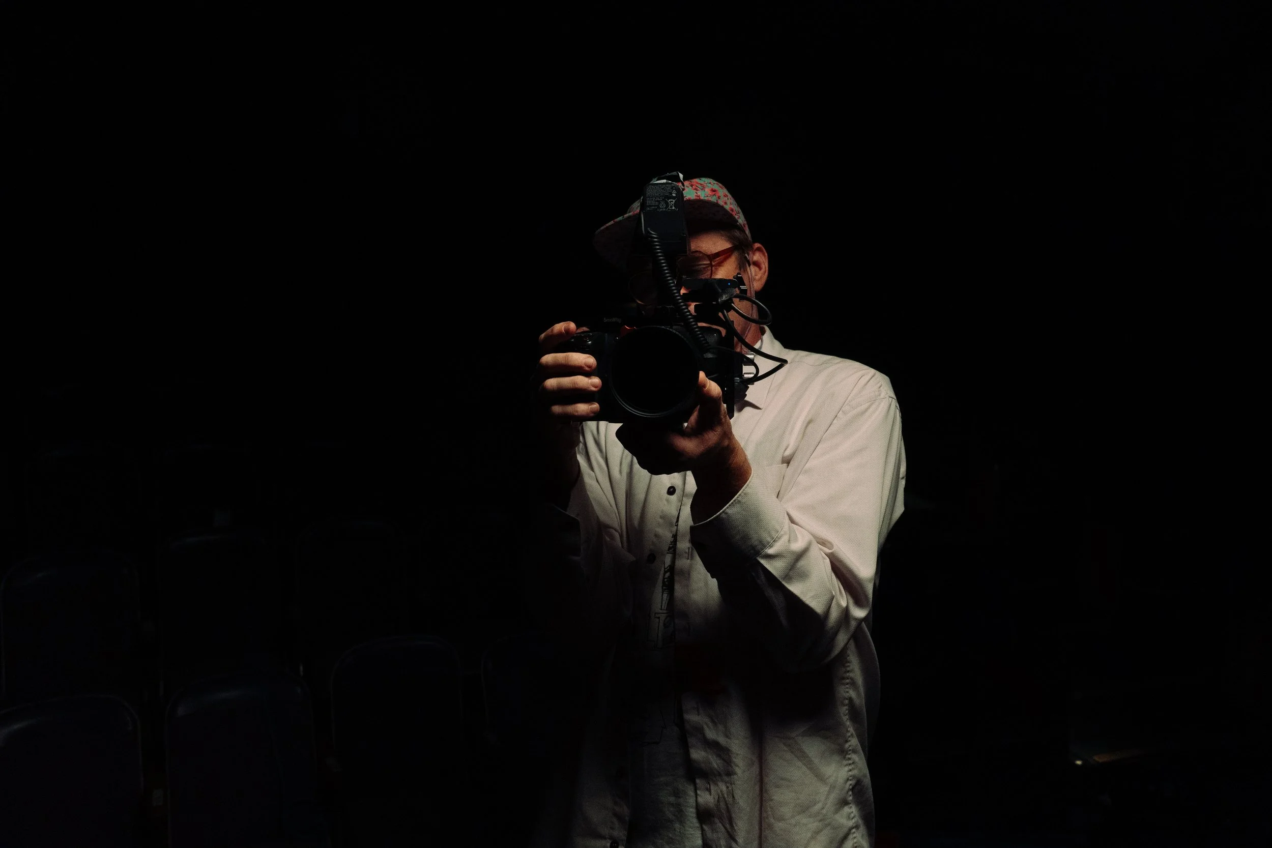 Person wearing glasses and a hat, taking a photo with a camera in a dark setting.