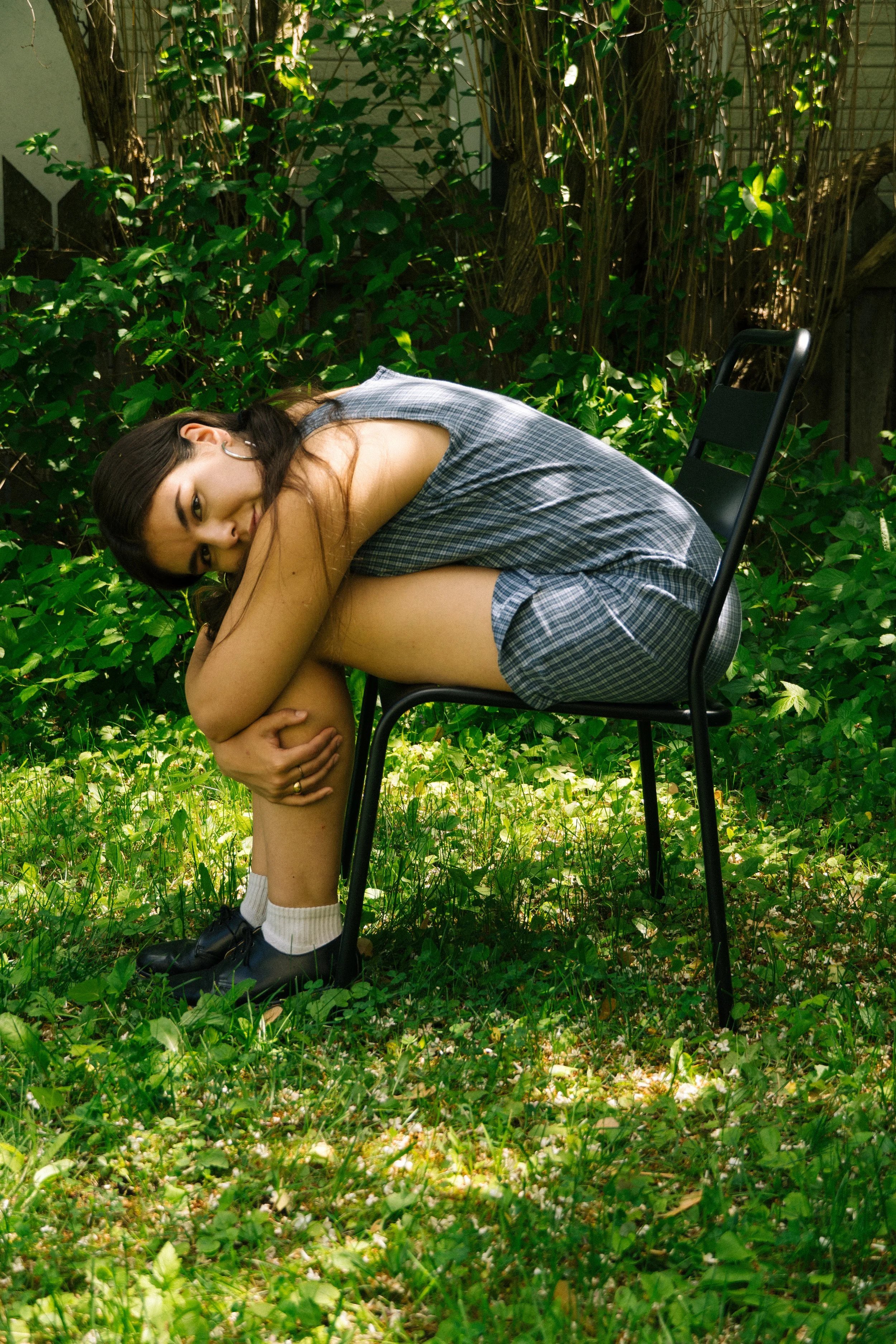 A young woman seated on a black chair outdoors among green foliage, wearing a plaid dress, black sneakers, and white socks, with dark hair and hoop earrings, smiling at the camera while holding her right knee with her arms, in sunlight.