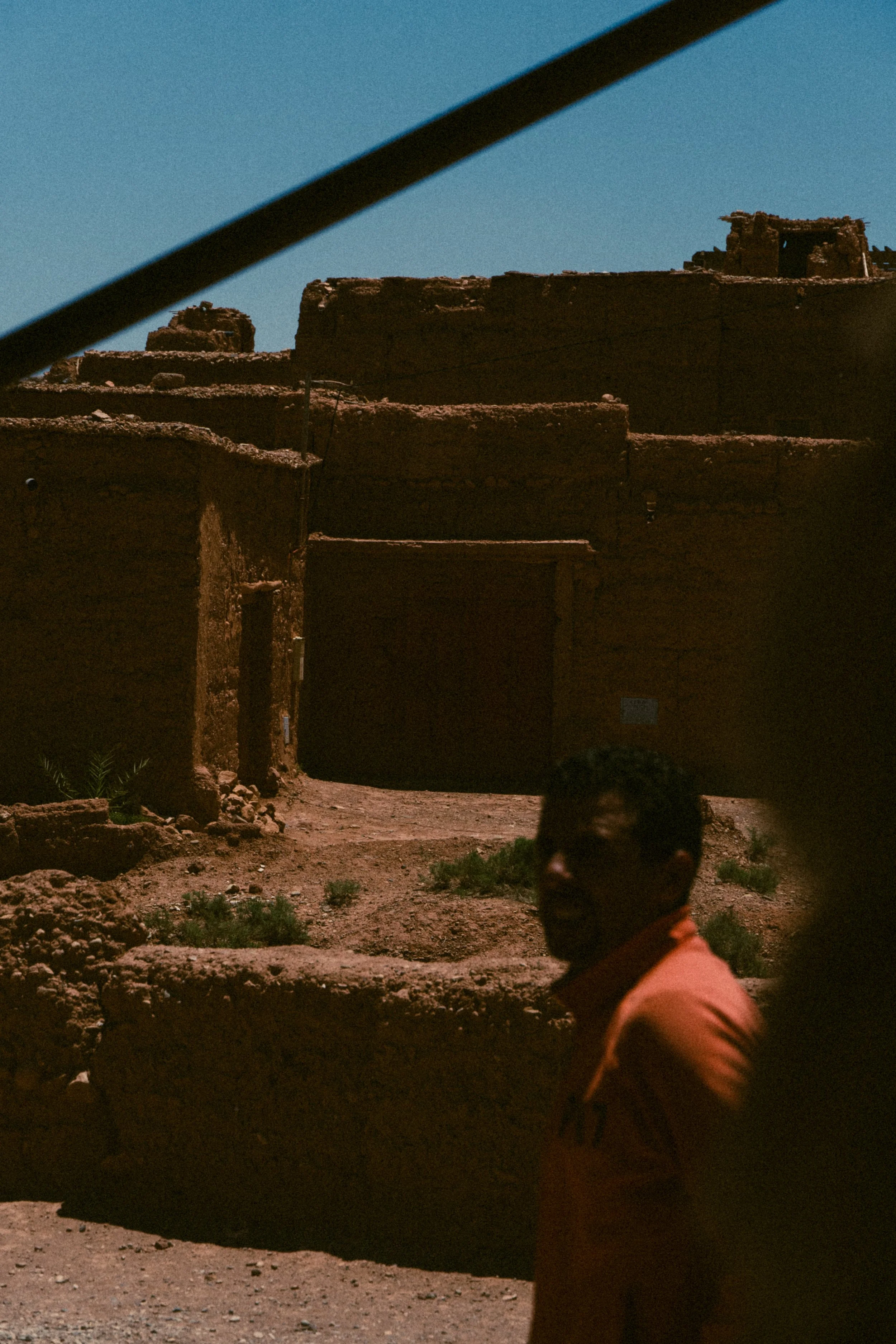 Person with glasses and orange shirt standing outdoors in front of an ancient mud-brick structure, under a clear blue sky.