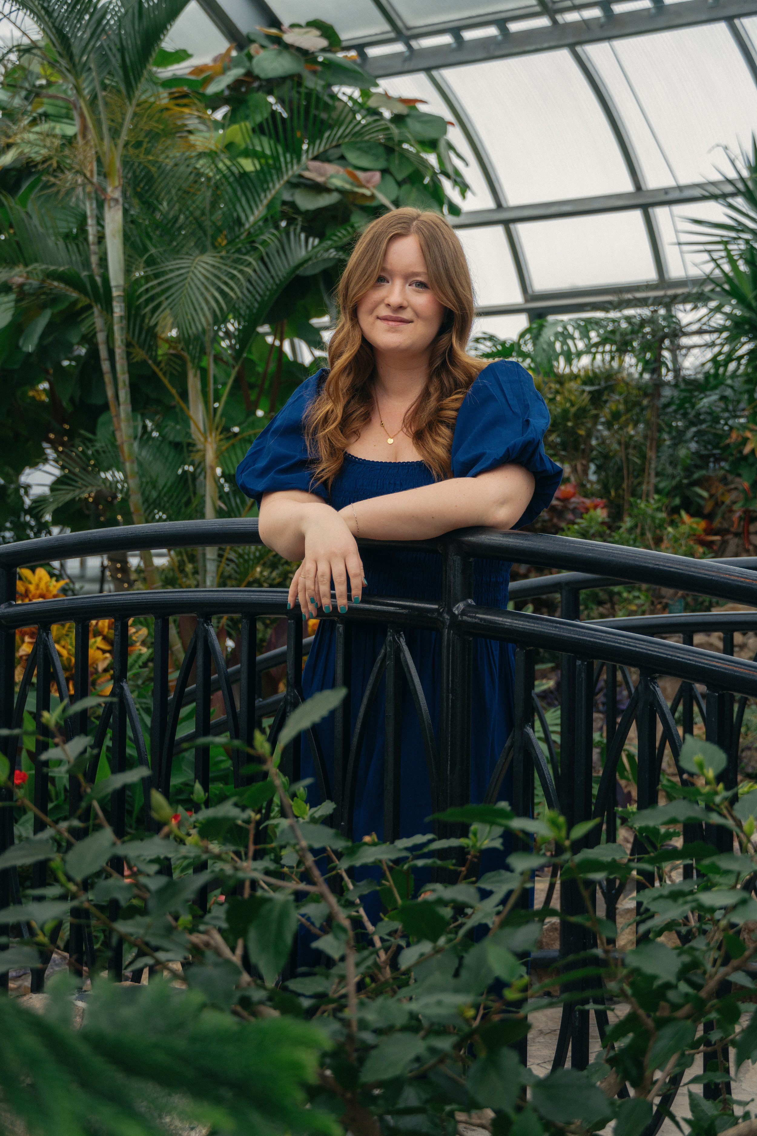 A woman with long, wavy, light brown hair and a smile, wearing a blue dress, standing in a lush greenhouse surrounded by various green plants and trees, with large windows in the background.