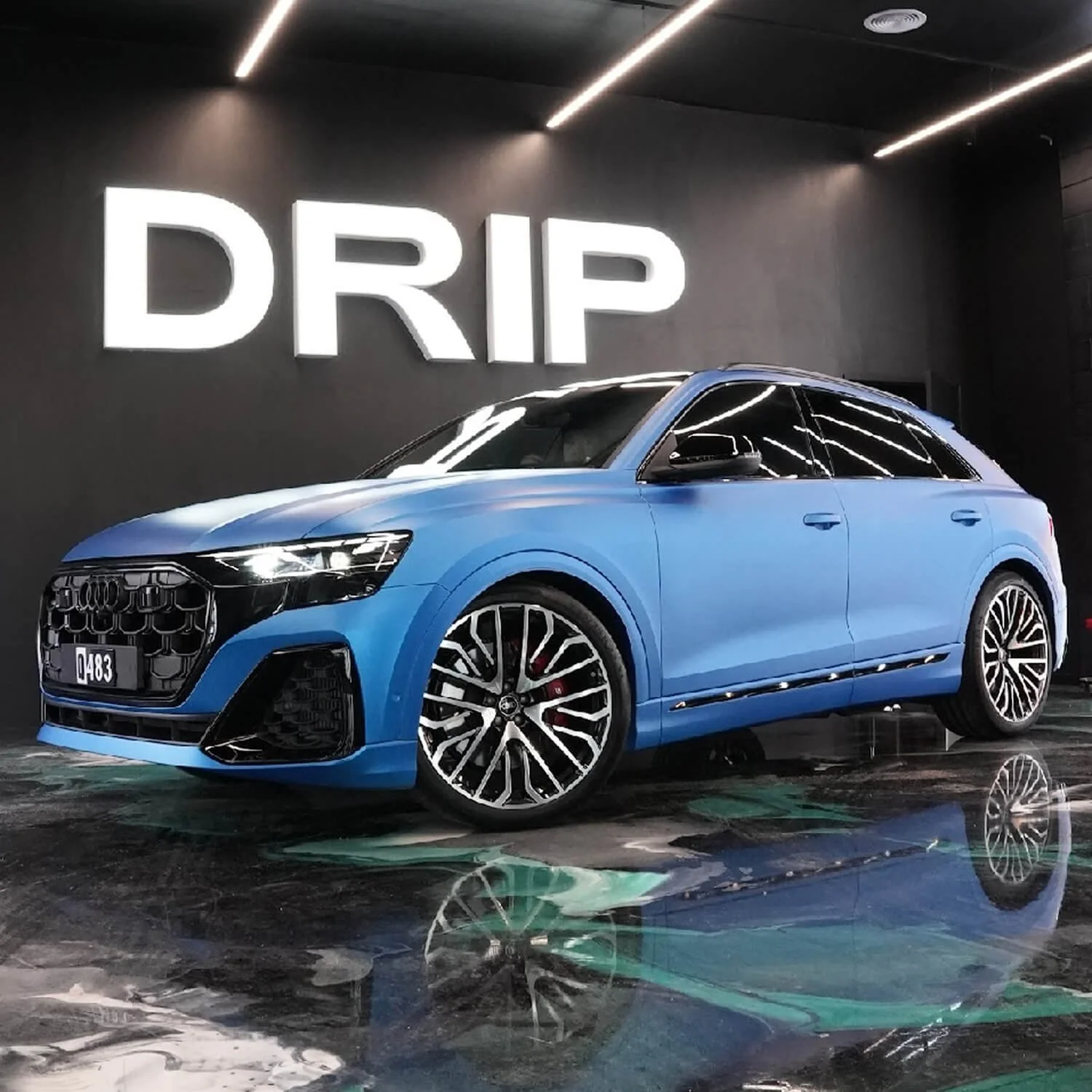 Blue luxury sedan car parked indoors, reflected on the shiny floor, with large illuminated 'DRIP' sign on a black wall behind it.