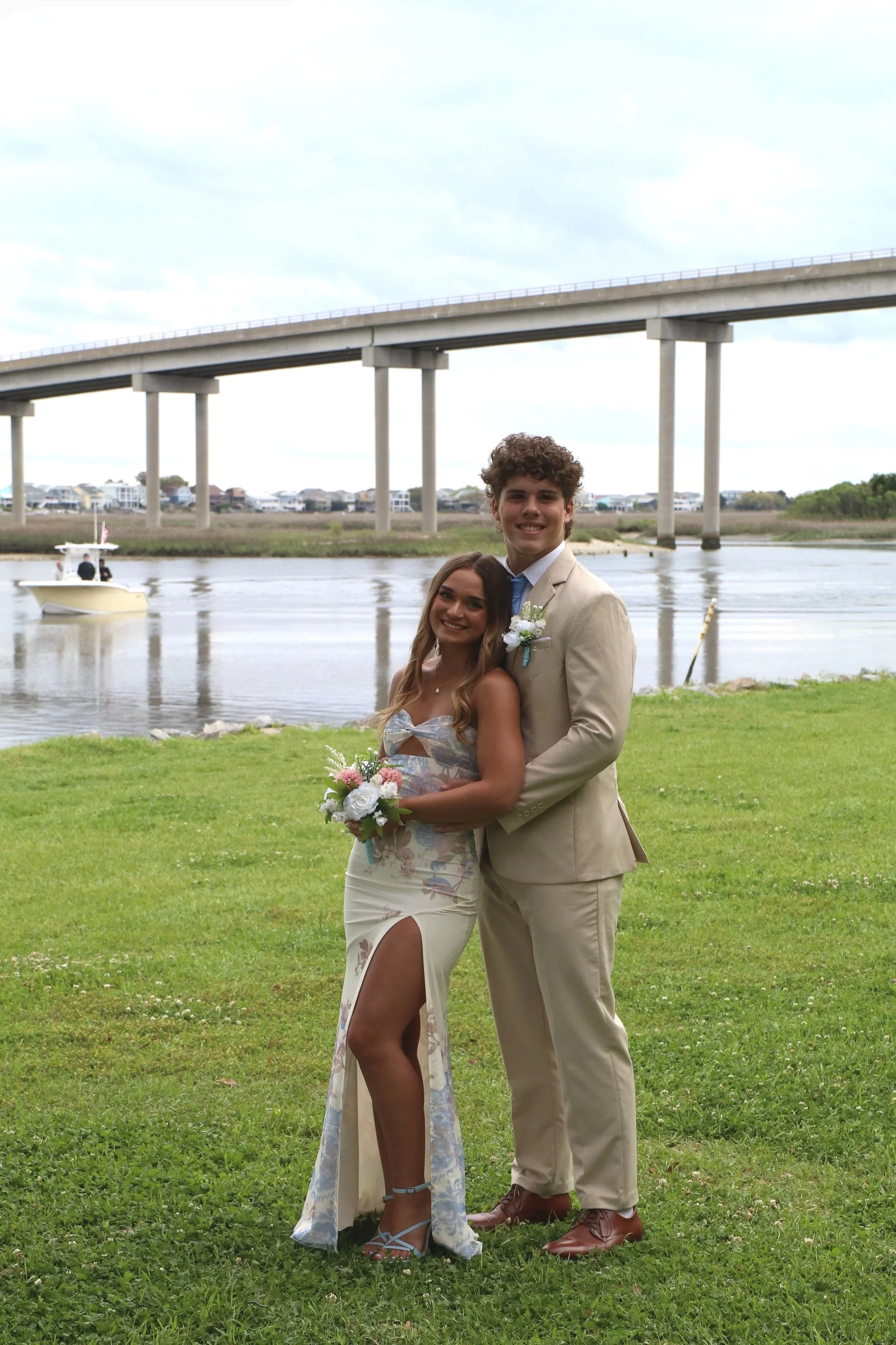 A young couple dressed in wedding attire standing on grass near a river with a bridge in the background. The woman is holding a bouquet and the man has a boutonniere.