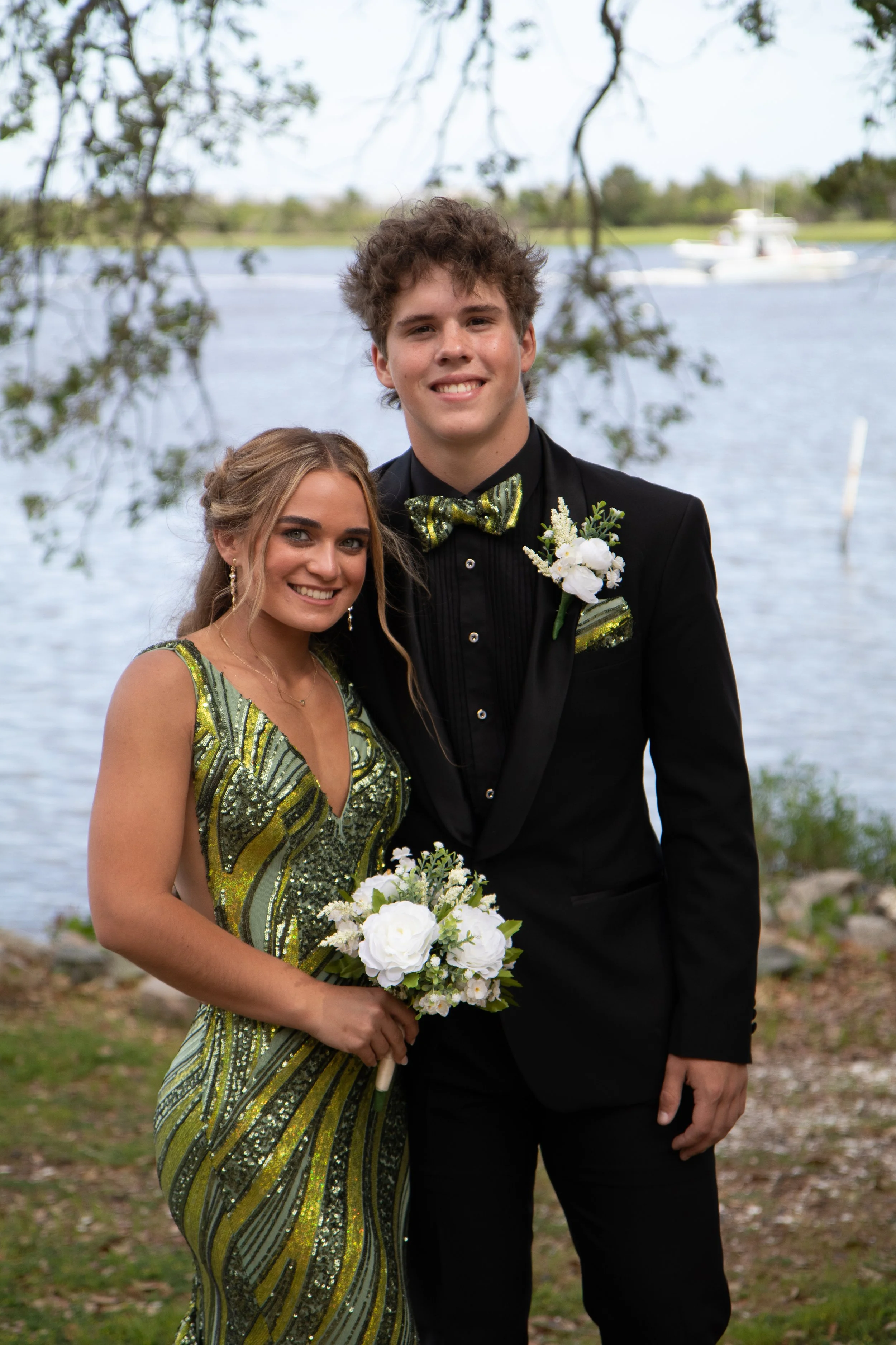 A young couple dressed in formal attire outdoors by a lake, with trees and a boat in the background. The woman wears a green and gold patterned dress, and the man wears a black tuxedo with a green and gold bow tie and boutonniere. They are smiling and holding bouquets of white flowers.