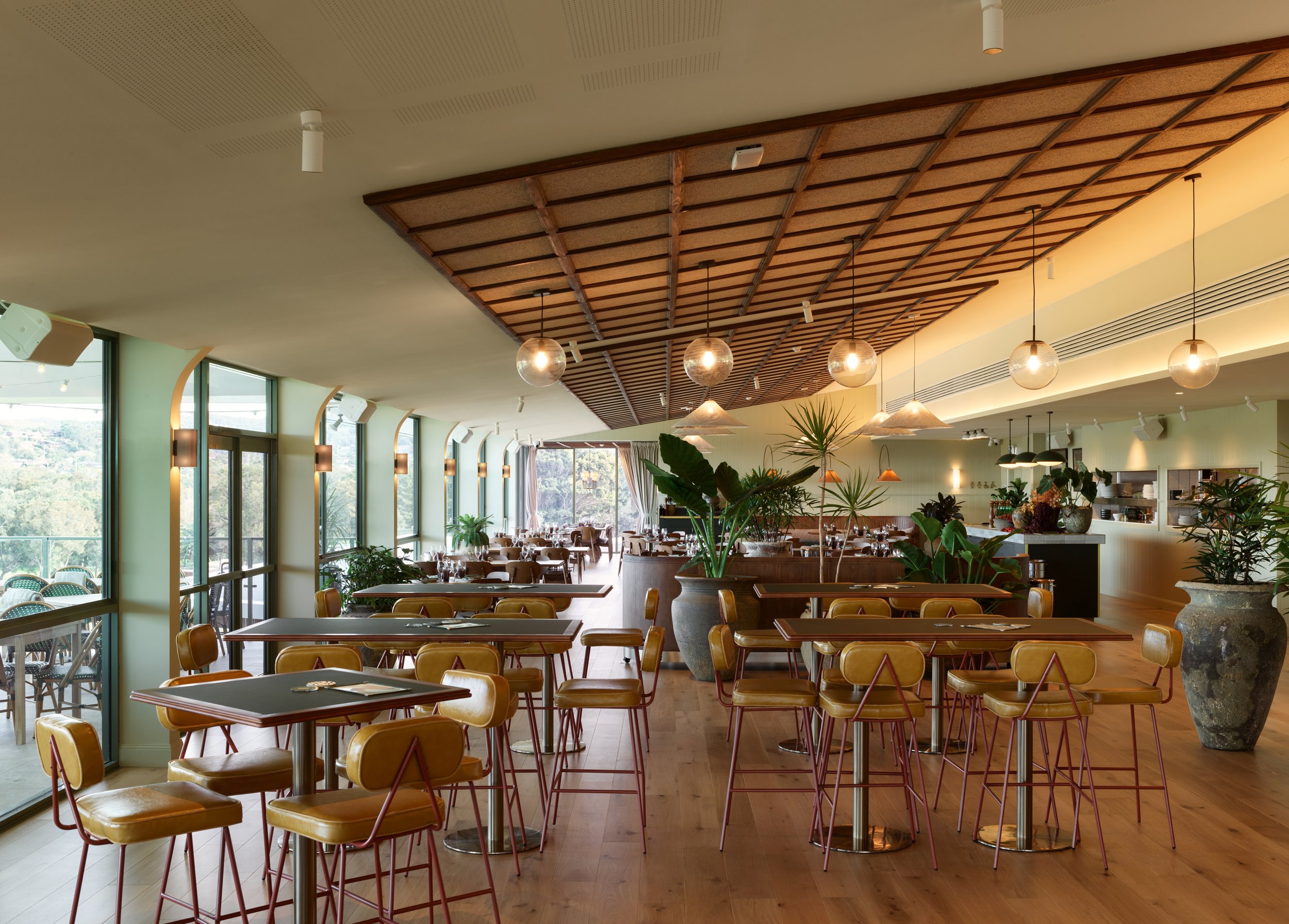 Interior of a modern restaurant or cafe with large windows, wooden flooring, and indoor plants. There are tables and chairs, with pendant lights hanging from a wooden ceiling feature.