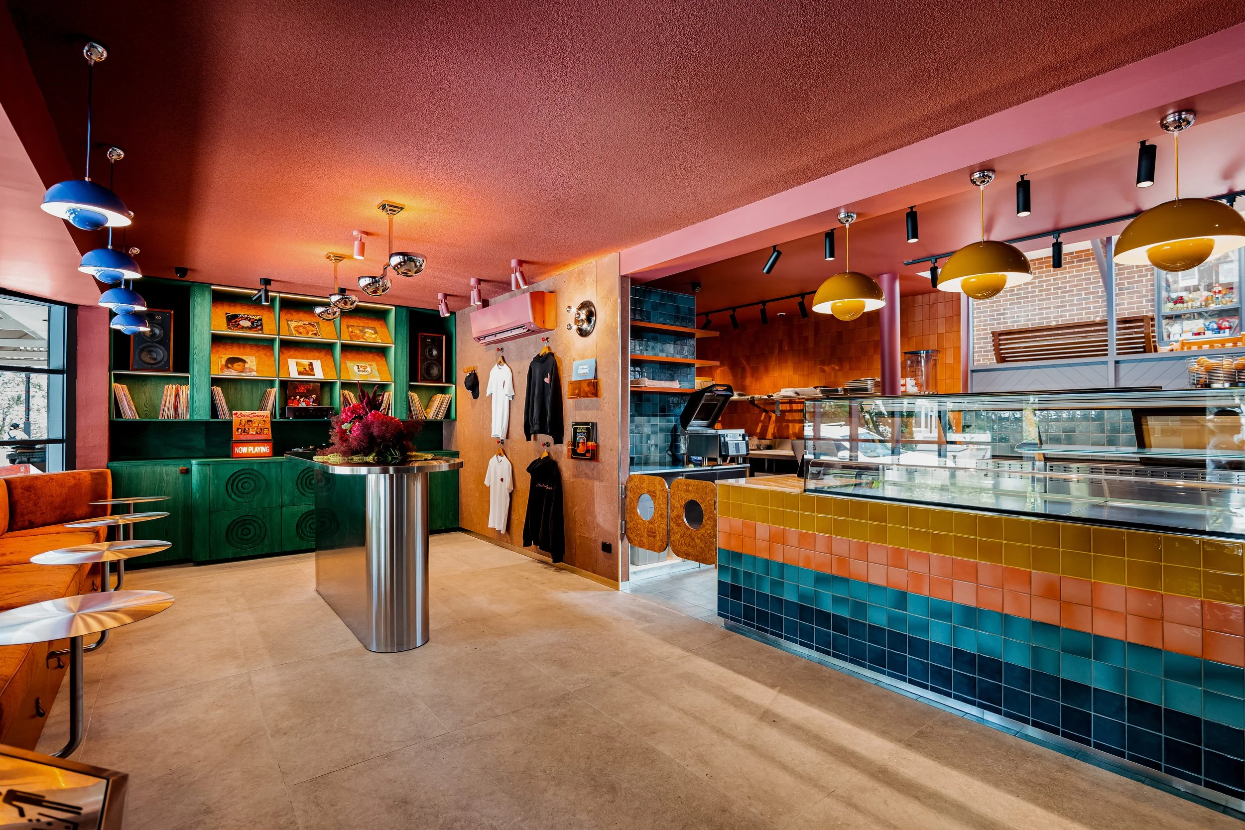 Interior of a colorful cafe with a green bookshelf, record player, vinyl records, and a seating area with orange chairs, a counter with a glass display, rainbow-colored tiles, and various decorative lighting.