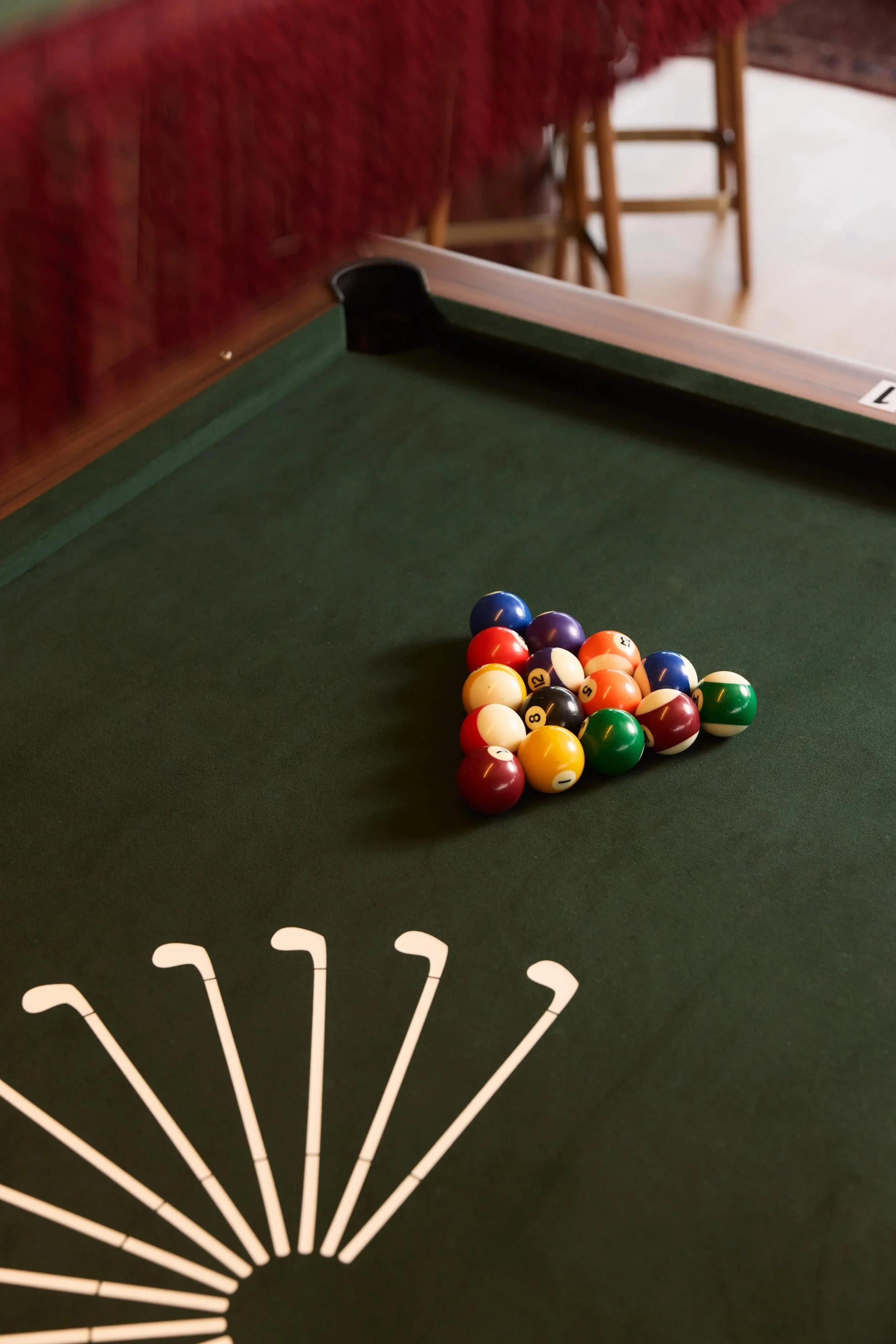 A pool table with a triangle of billiard balls arranged at the rack line, including solid and striped balls. The green felt surface of the table is visible, with a white painted semi-circle design and a corner pocket. In the background, part of a wooden chair and a stooled chair are visible.
