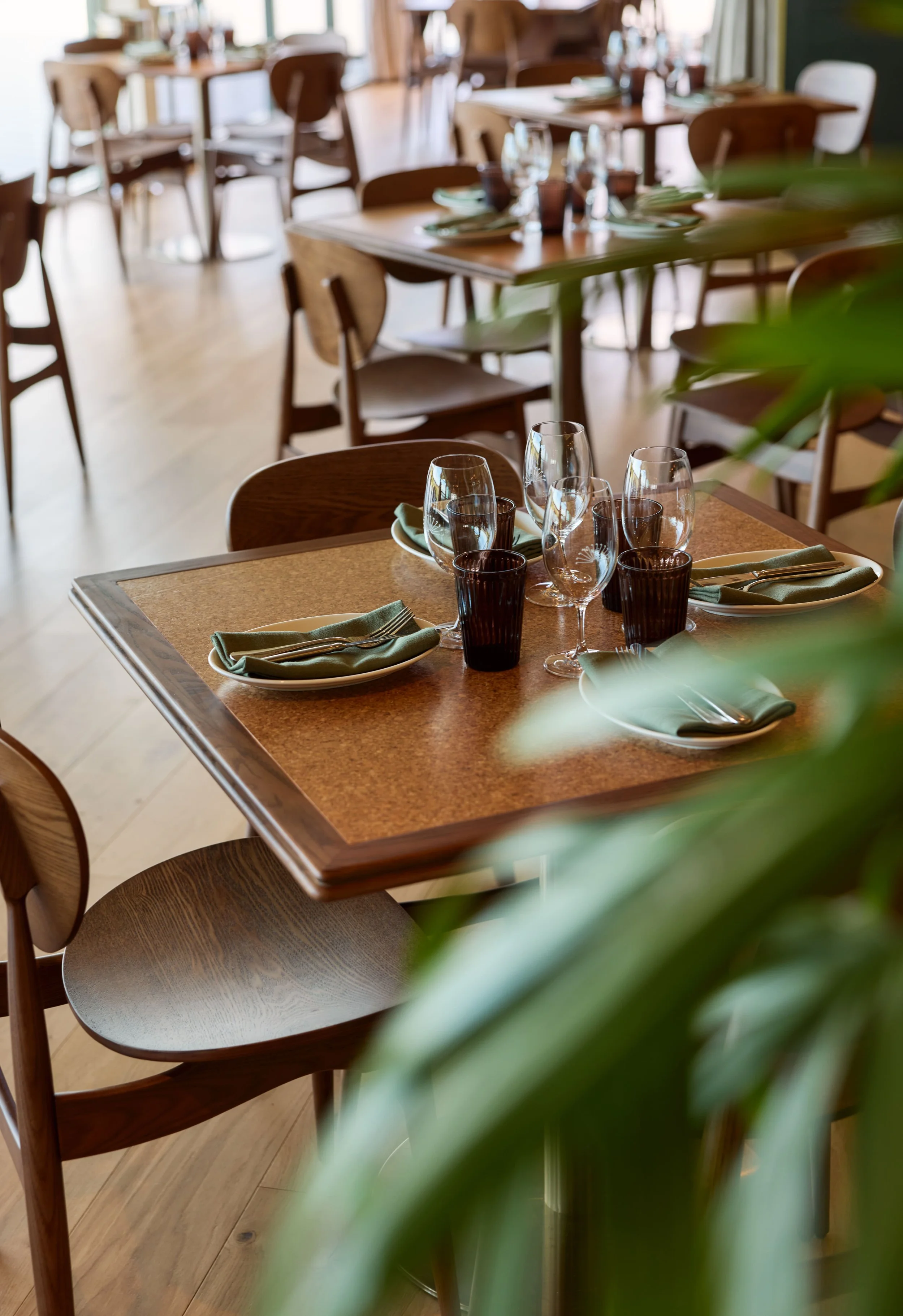 A dining table set with green napkins, glasses, and black cups in a restaurant with multiple wooden tables and chairs, partially obscured by green plant leaves in the foreground.