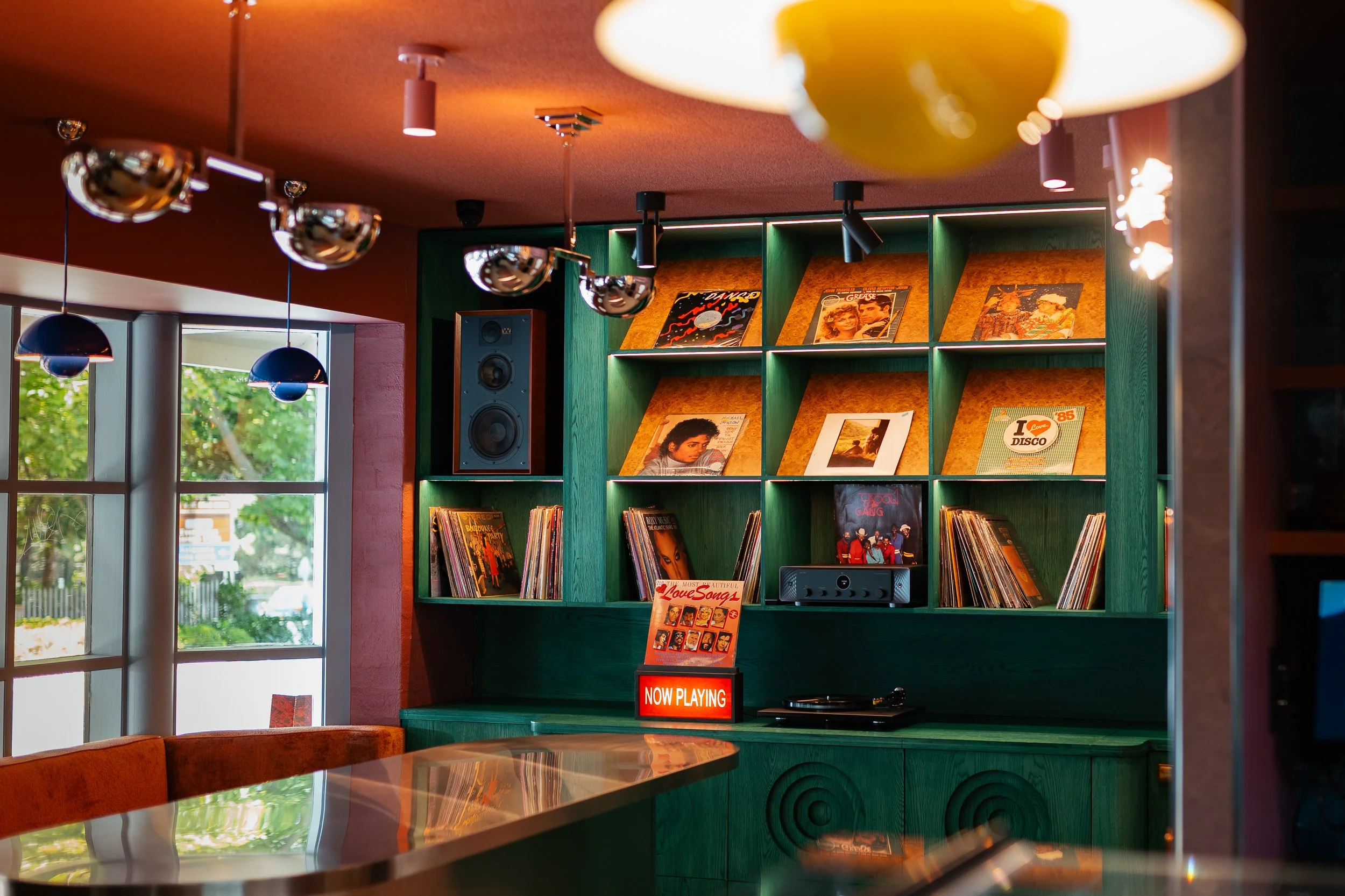 Indoor scene of a record store or listening lounge with a green shelving unit displaying vinyl records and posters, a window with greenery outside, hanging ceiling lights, and a sign that says 'Now Playing.'